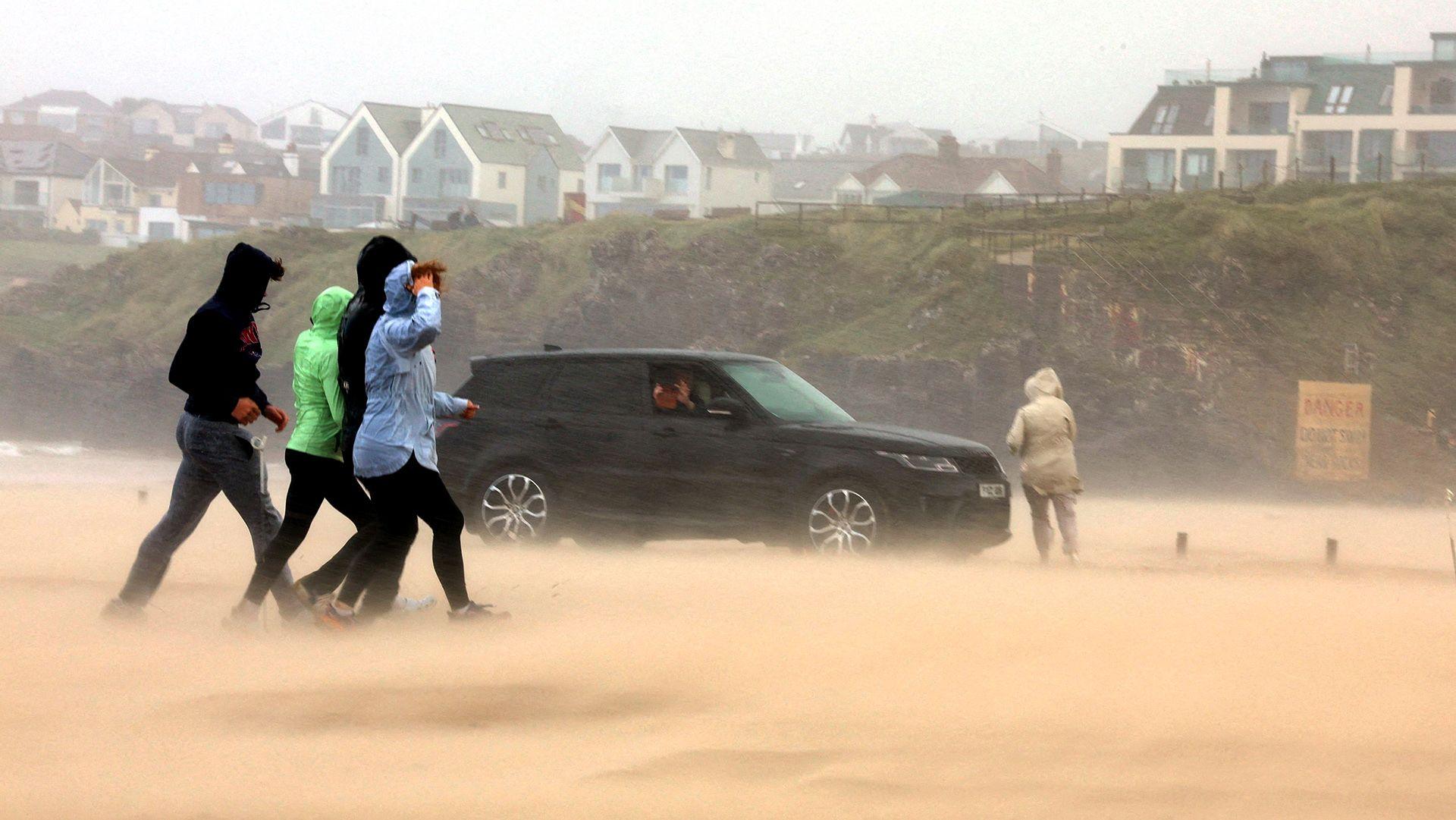 A group of four people walk across sand which is being blown up by strong wind. A black car is in the distance