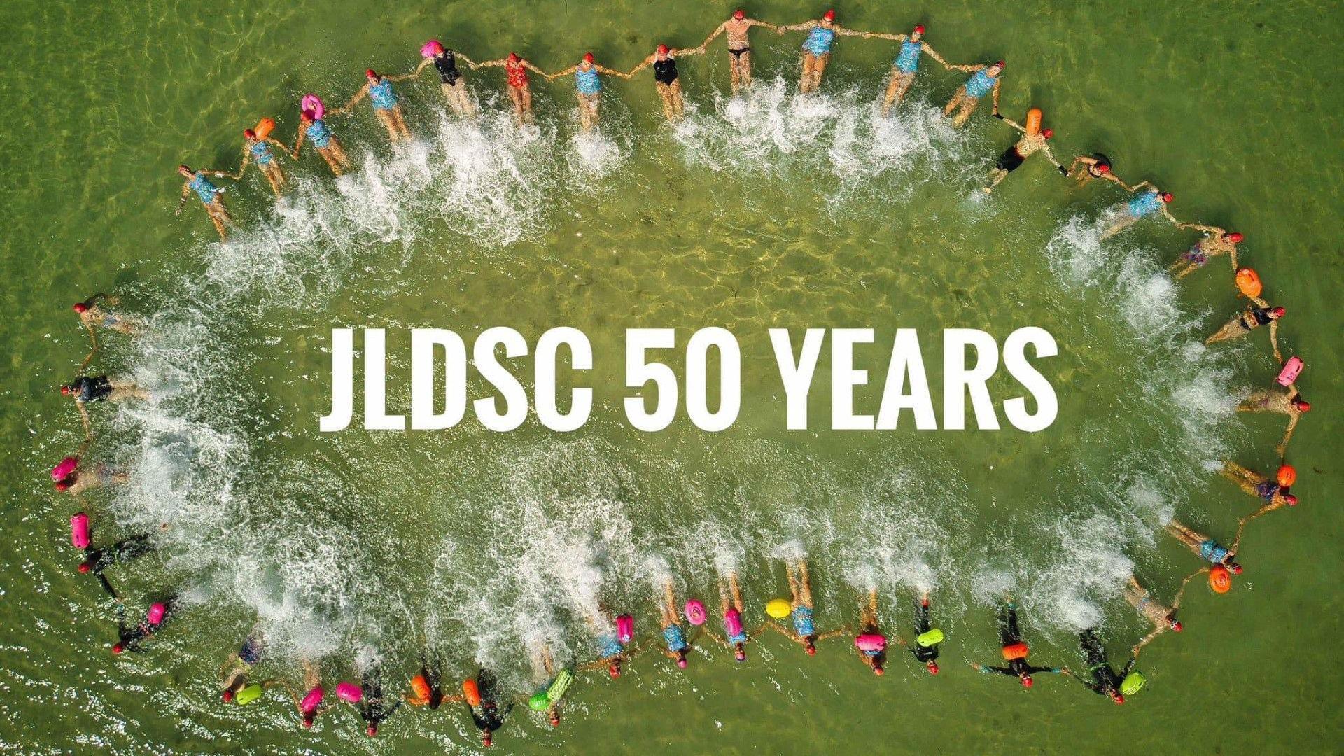 An aerial photo looking down into the sea. The water has a green tinge. Multiple members of the Jersey Long Distance Swimming Club are floating in the water in a long circle shape, all holding hands. In the centre of the circle the text 'JLDSC 50 YEARS' has been edited onto the image.