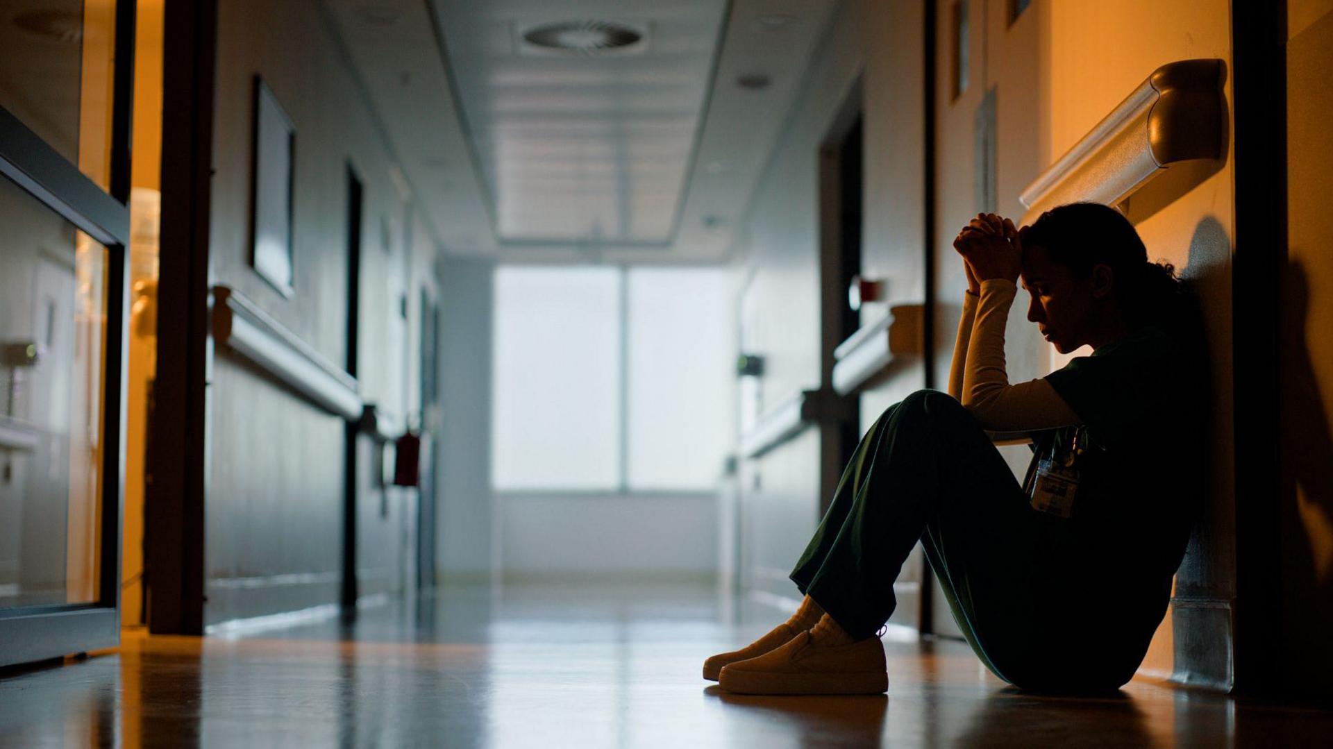 A female staff worker sits on the floor of a hospital corridor with her head in her hands. She is in the shadows