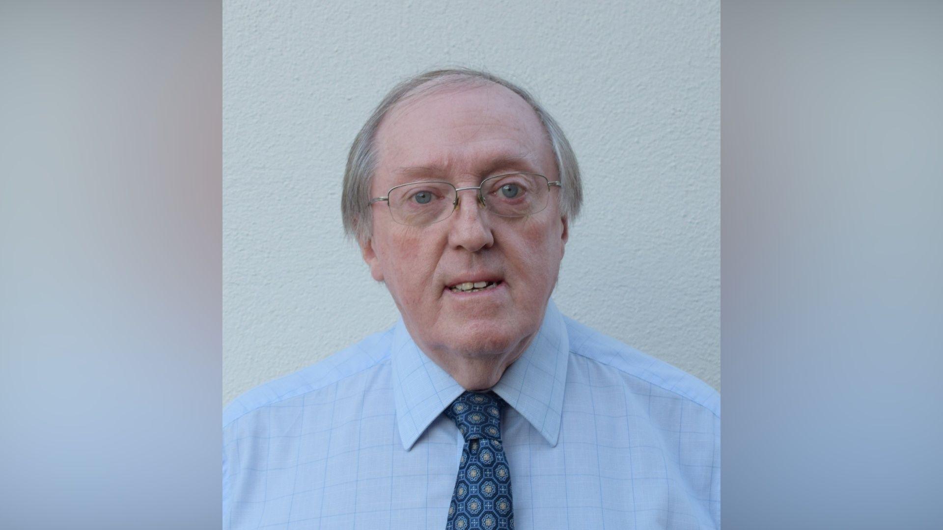 Prof John Sweeney has thinning grey hair and wire framed glasses. He is wearing a light coloured shirt and a spotted tie