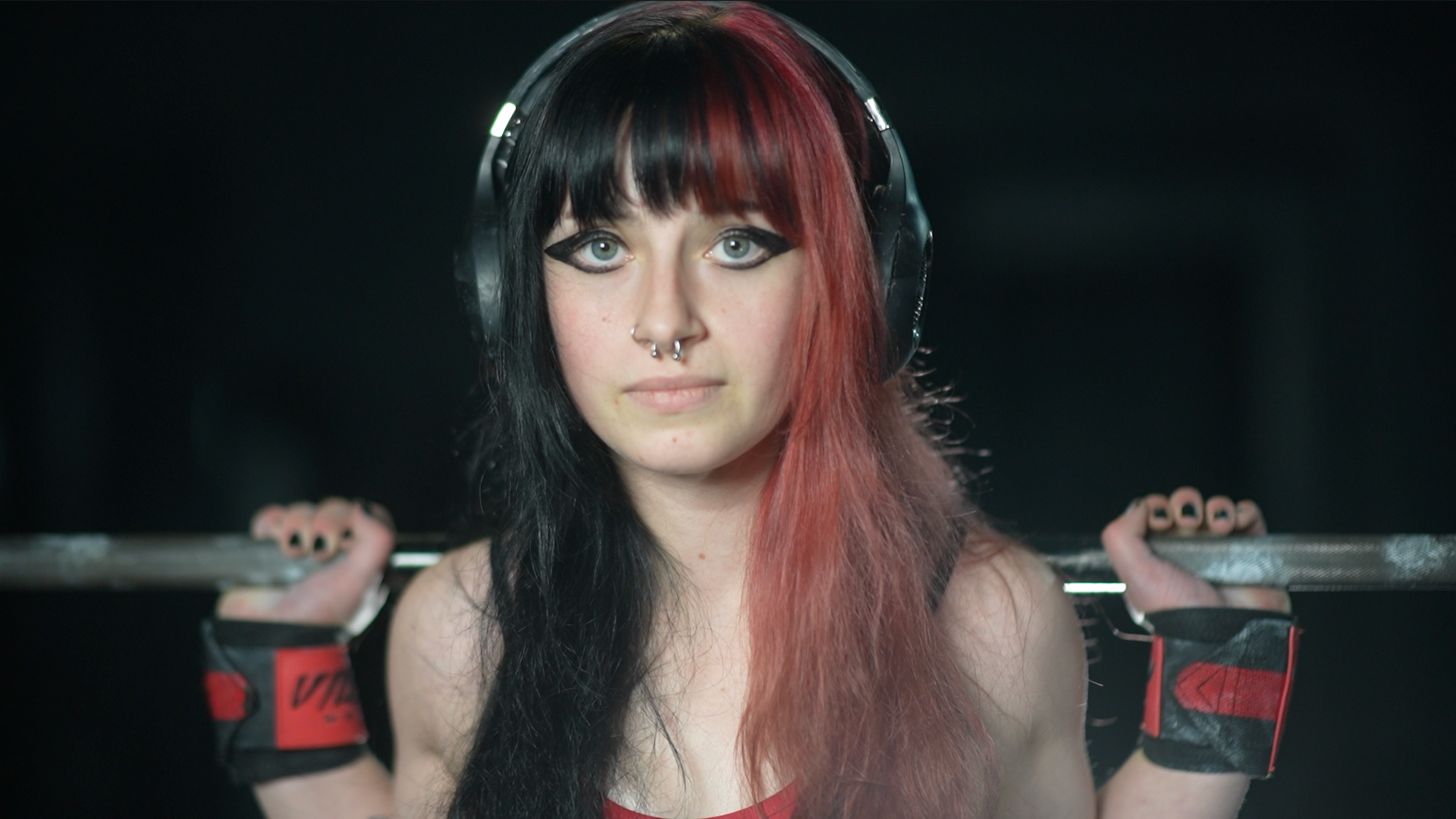 A photo of Miss Churchyard looking directly at the camera. She is half smiling and has a squat barbell sitting across her shoulders. She is holding the barbell and has wrist straps on each arm. 