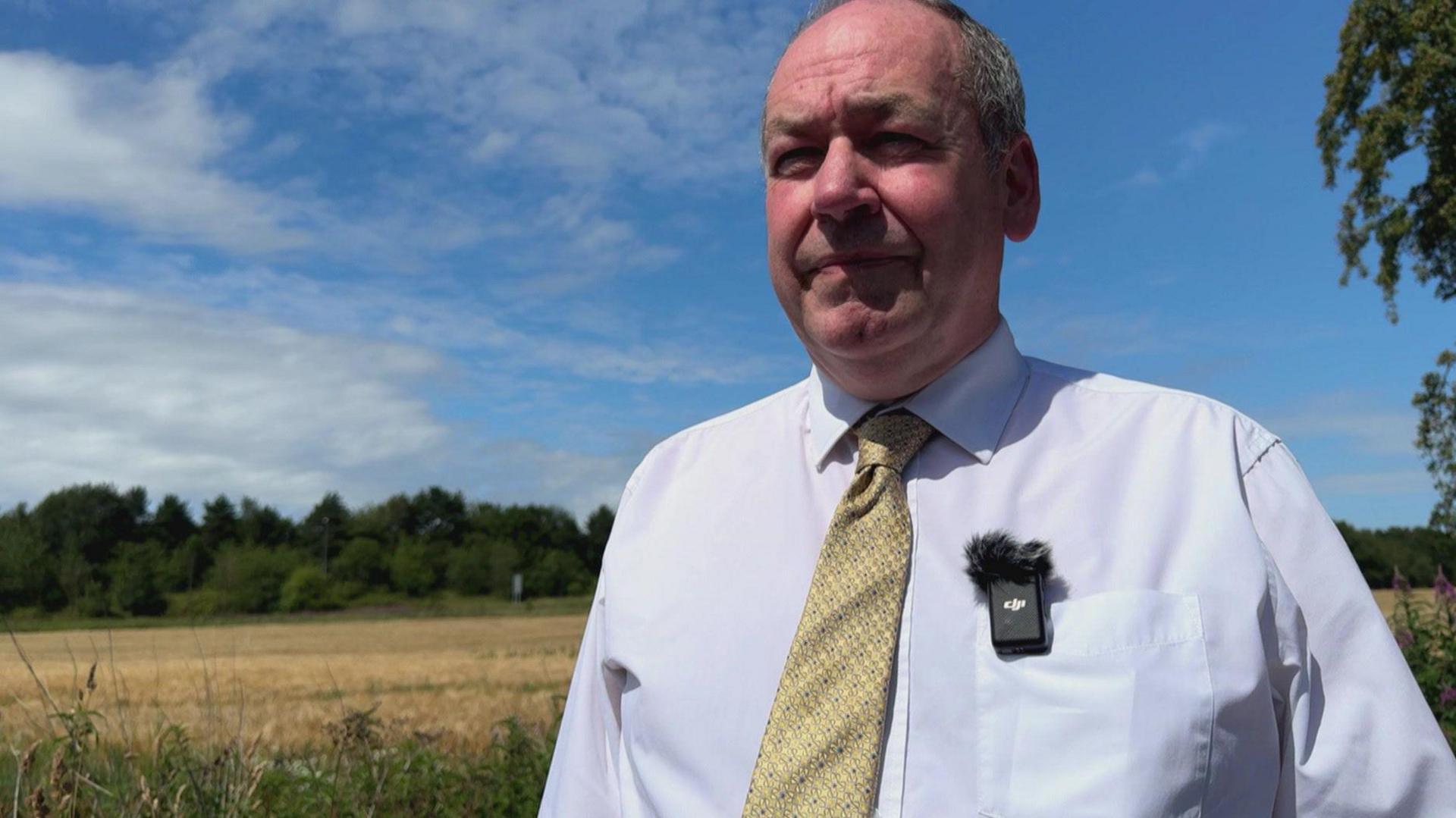 An image of Adrian Owens standing in front of a yellow field, with Rainford Road in the background. The background is tree-lined. The sky is blue with scattered clouds. Councillor Owens is wearing a white shirt with a gold tie and has a microphone clipped to his right pocket.
