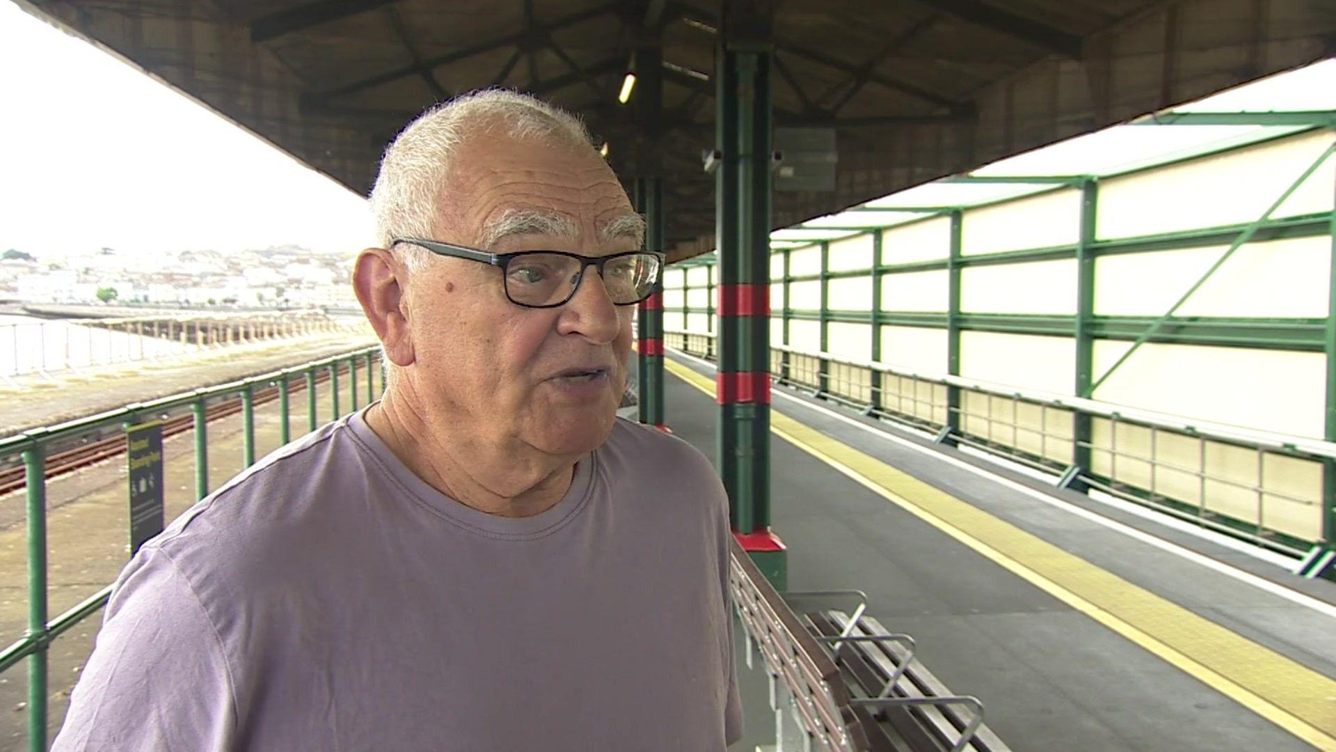 A man with short grey hair and glasses, and wearing a purple t-shirt, stands on a railway platform with the railway behind him. Behind the railway is the sea with the pier running across it, with the mainland just about in view.