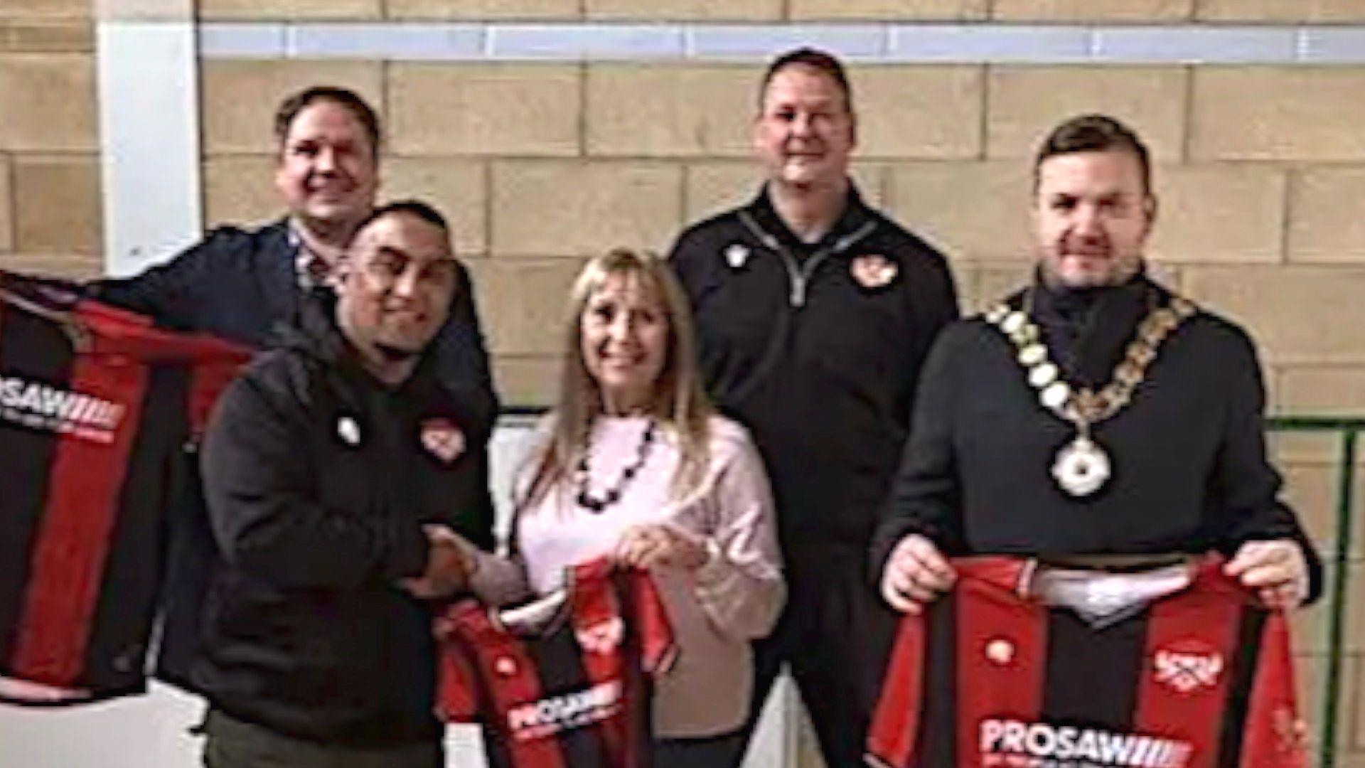 Four men, all dressed in black tops, and a woman, in a pink jumper, stand in a sports hall holding up three red and black football shirts.