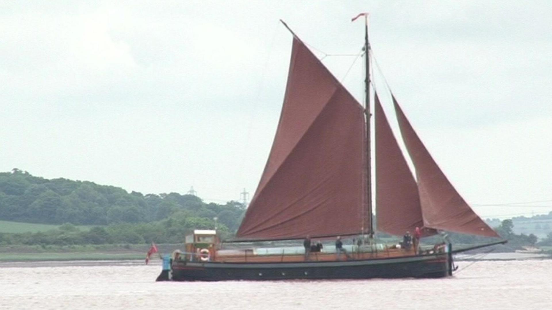 A black boat with a large red sail on the Humber. In the background is a tree-covered bank. 