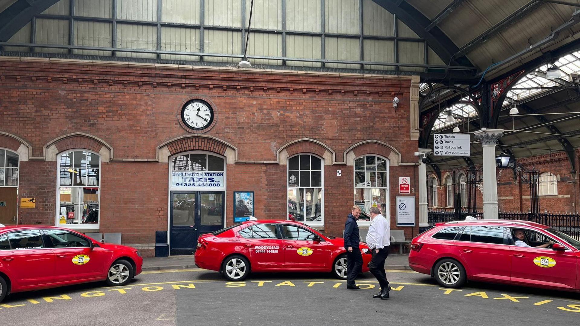 Three red taxis are parked outside of Darlington station at the taxi rank. Two men are stood beside the taxis speaking.