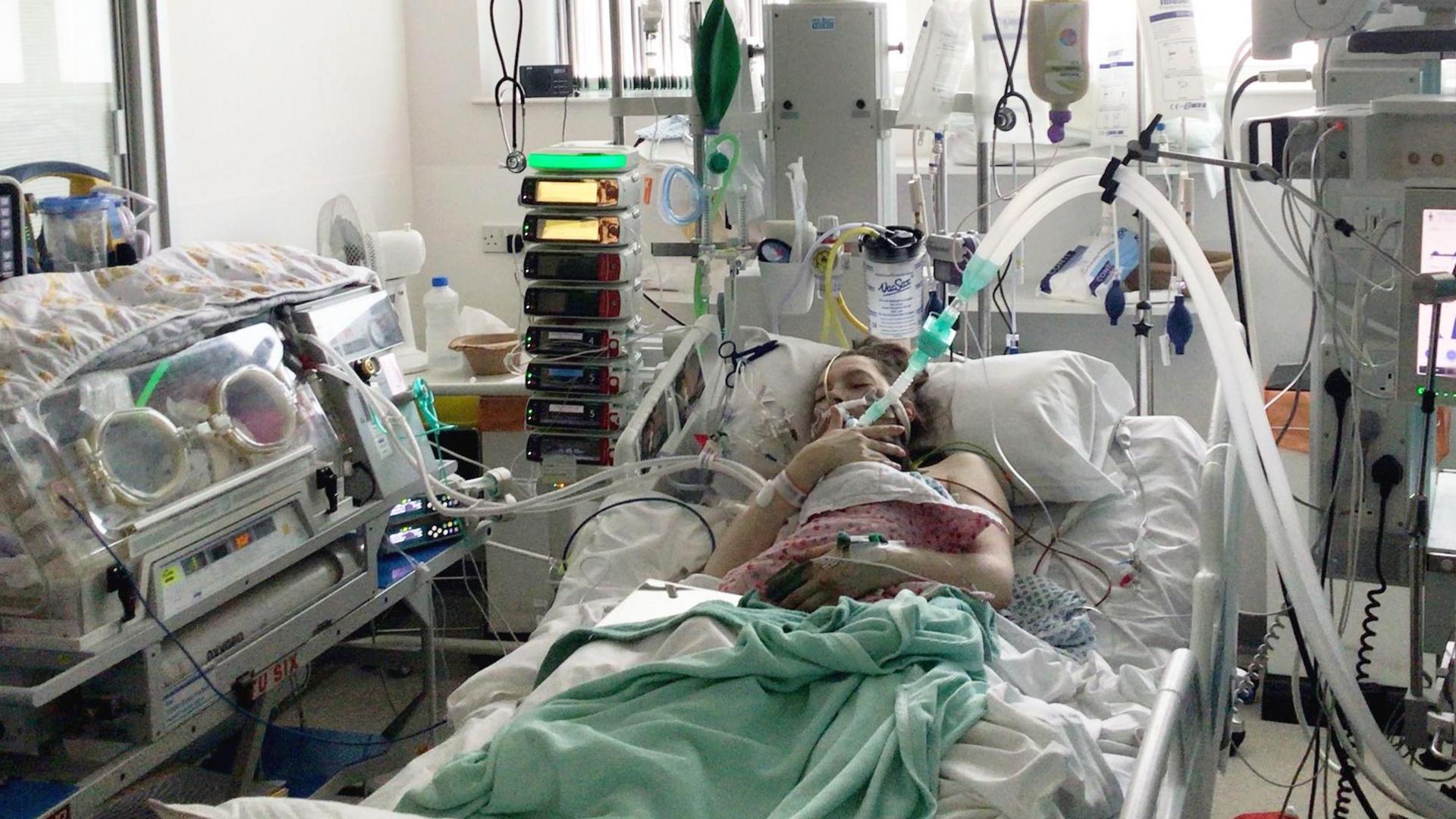 Helen holding baby Maeve. She has a large tube in her mouth and wires attached all over her body. There is a baby incubator to the left hand side