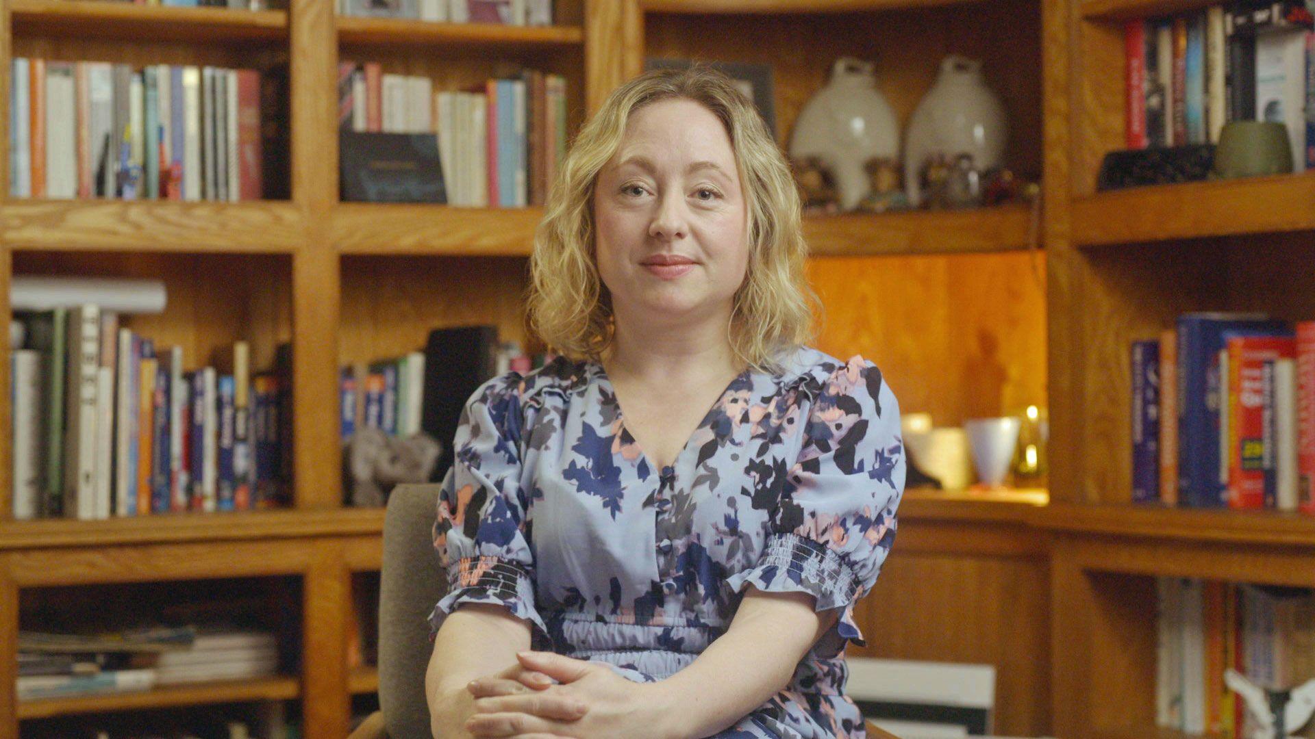 A grab from the TV programme of journalist Catriona Stewart. She is sitting in a study room with wooden bookshelves behind her. She has her hands clasped in her lap as she looks at the camera. She is wearing a lavender-patterned dress.
