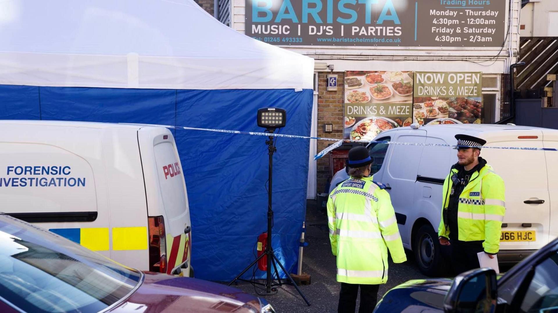 Two police officers in yellow high-vis jackets standing in front of a white van parked in front of a food outlet called Barista. To its left is a large blue and white police forensic tent and in front of that is a white police forensics van