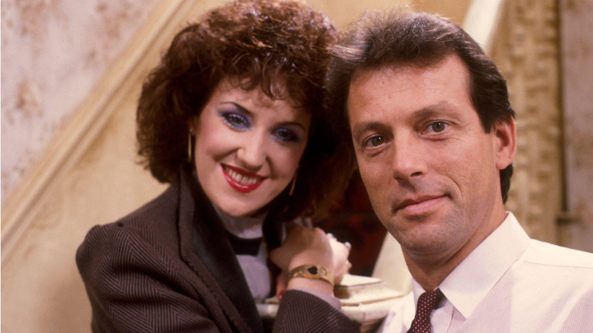 anita dobson and leslie grantham posing around the banister of the staircase behind the queen vic bar