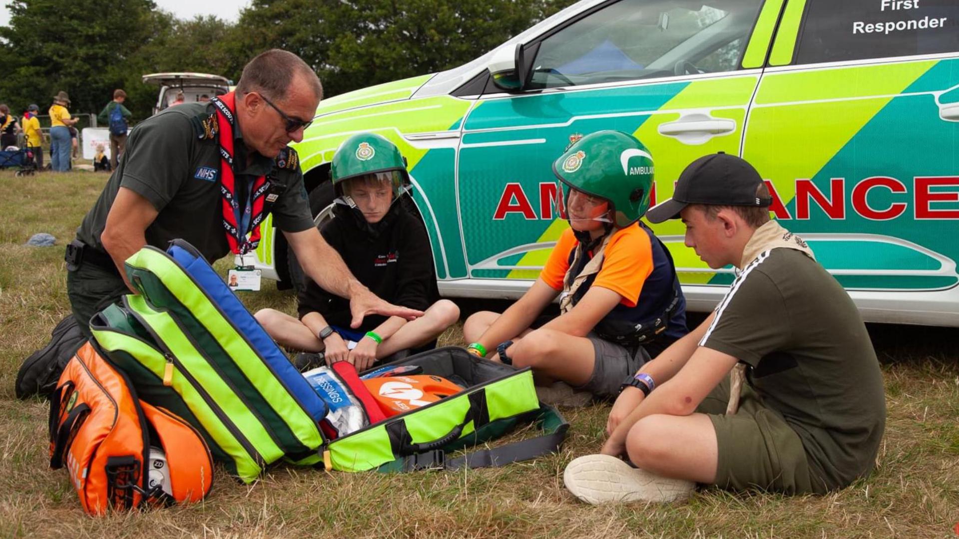 Stuart Little kneeling in front of three children on a field while showing them life-saving equipment, held in an open green bag. He has his hand over the bag and the children are all watching what he is doing. They are in front of a community first responder ambulance, which is an estate car with fluorescent branding across it.