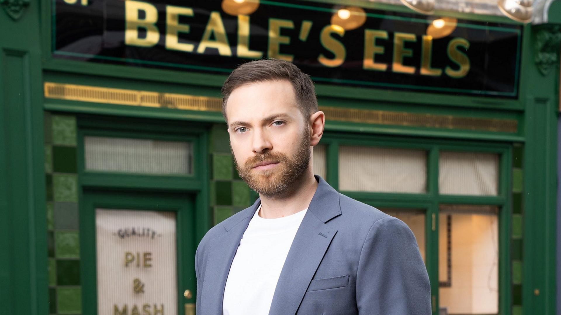 A photo shows actor Matt Di Angelo standing in front of a dark green fronted shop. It is called Beale's Eels and is a Pie and Mash shop in the BBC show Eastenders. 