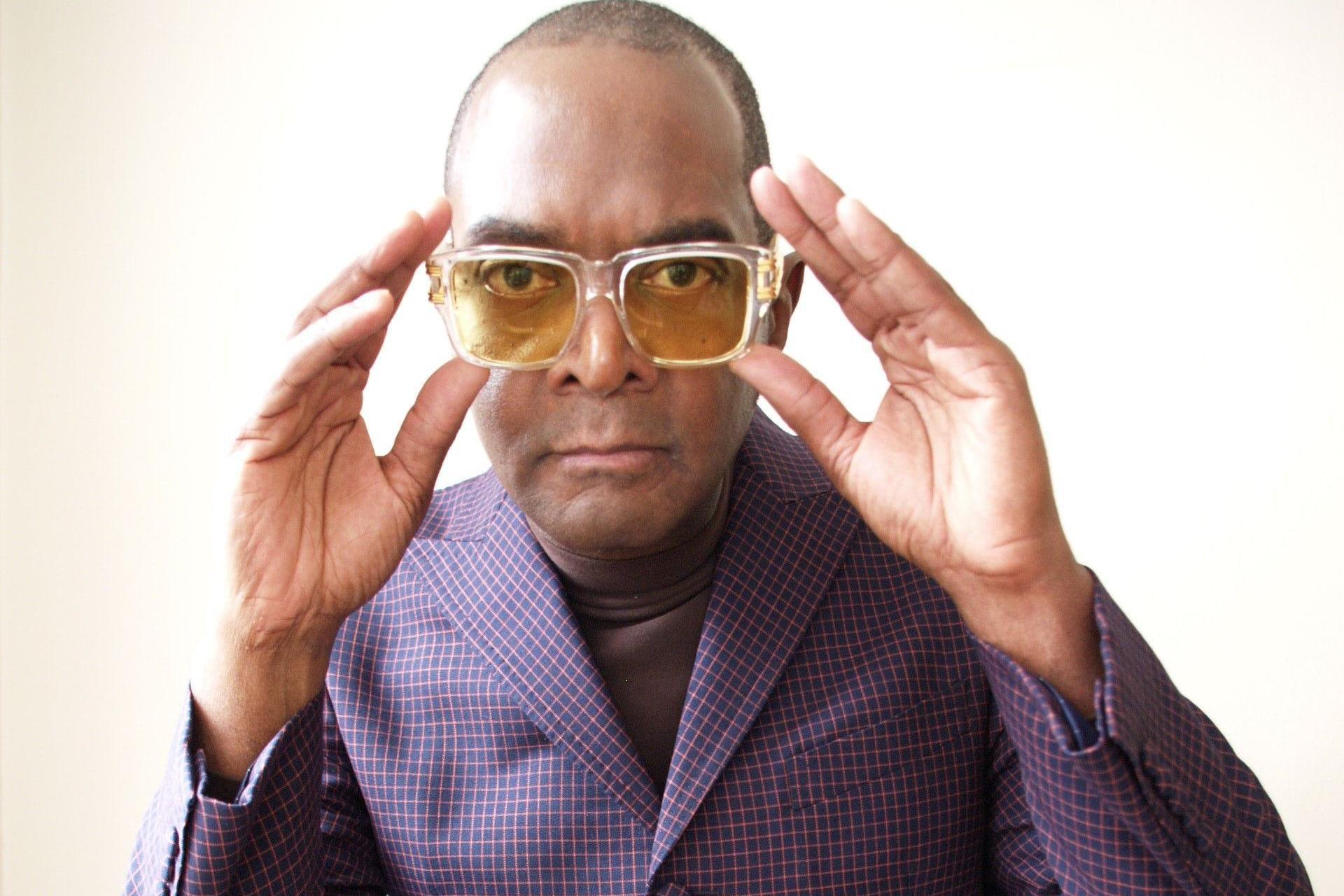 Dennis Morris wearing a blue/purple suit with pink lines in it. He is wearing yellow tinted glasses which he is holding with both hands