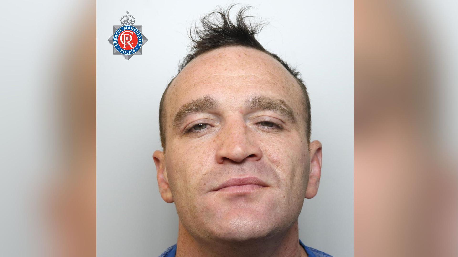 Kyle Woods is looking directly at the camera in this police photo. He looks like he is smiling slightly. He has blue eyes and short brown hair styled into a quiff. There is a Greater Manchester Police logo on the top left of the photo.