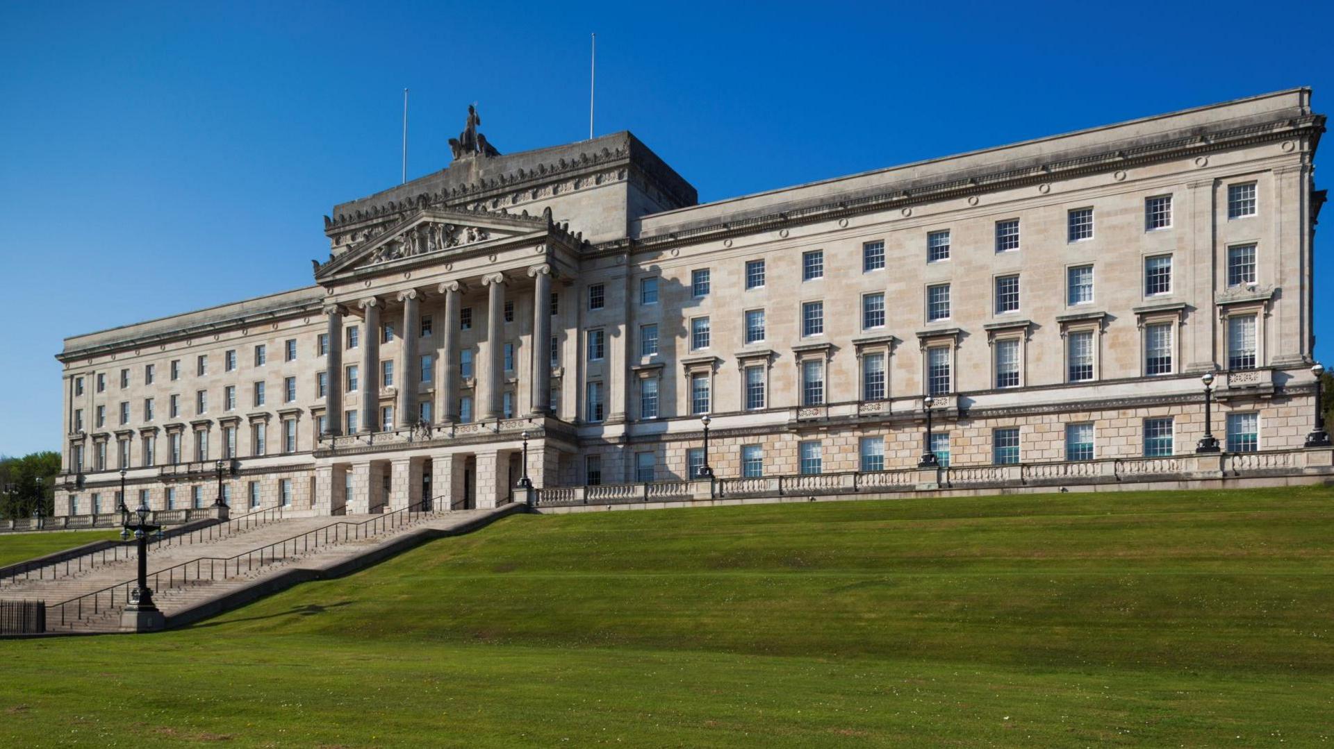 A image of Stormont's parliament buildings against a blue sky. The building is a large, white stone building with a number of windows and six large columns at the front of it.