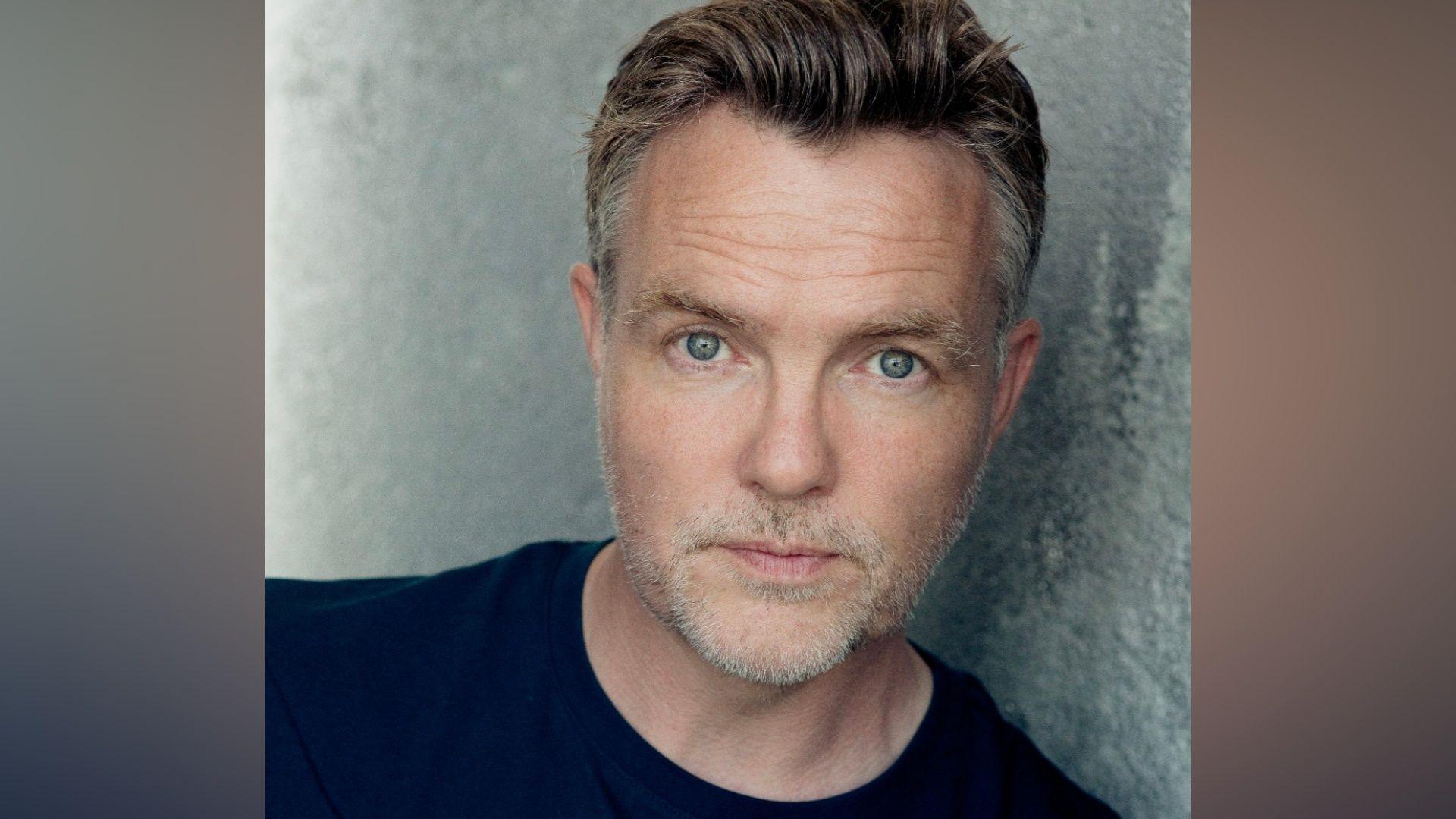 A man with blue eyes stares into the camera. He is wearing a navy top. He has brown hair swept and style back with shorter grey hairs at the side. He has grey stubble.