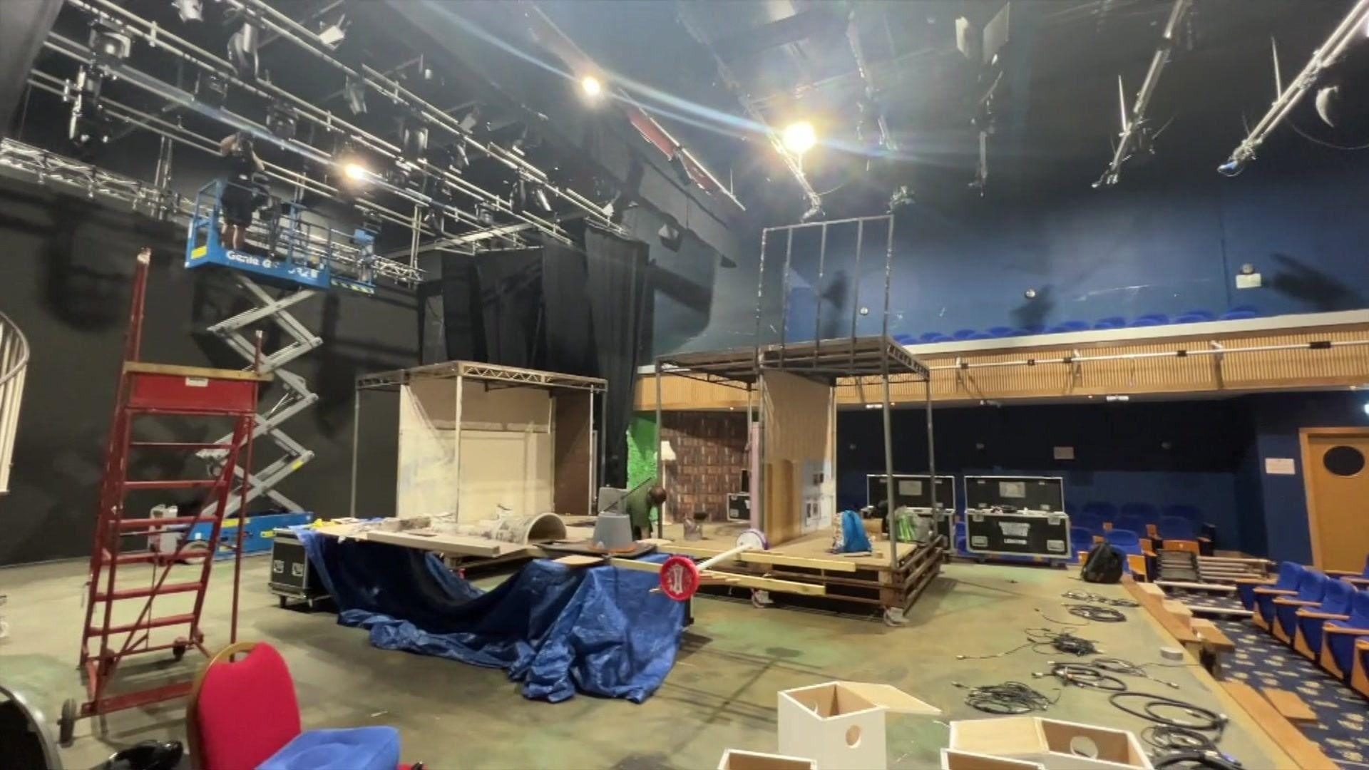 The stage of the theatre with scaffolding and half-built set on it. There are silver poles with stage lighting hanging from them on the ceiling, with a man carrying out some maintenance to one of them using a scissor lift.