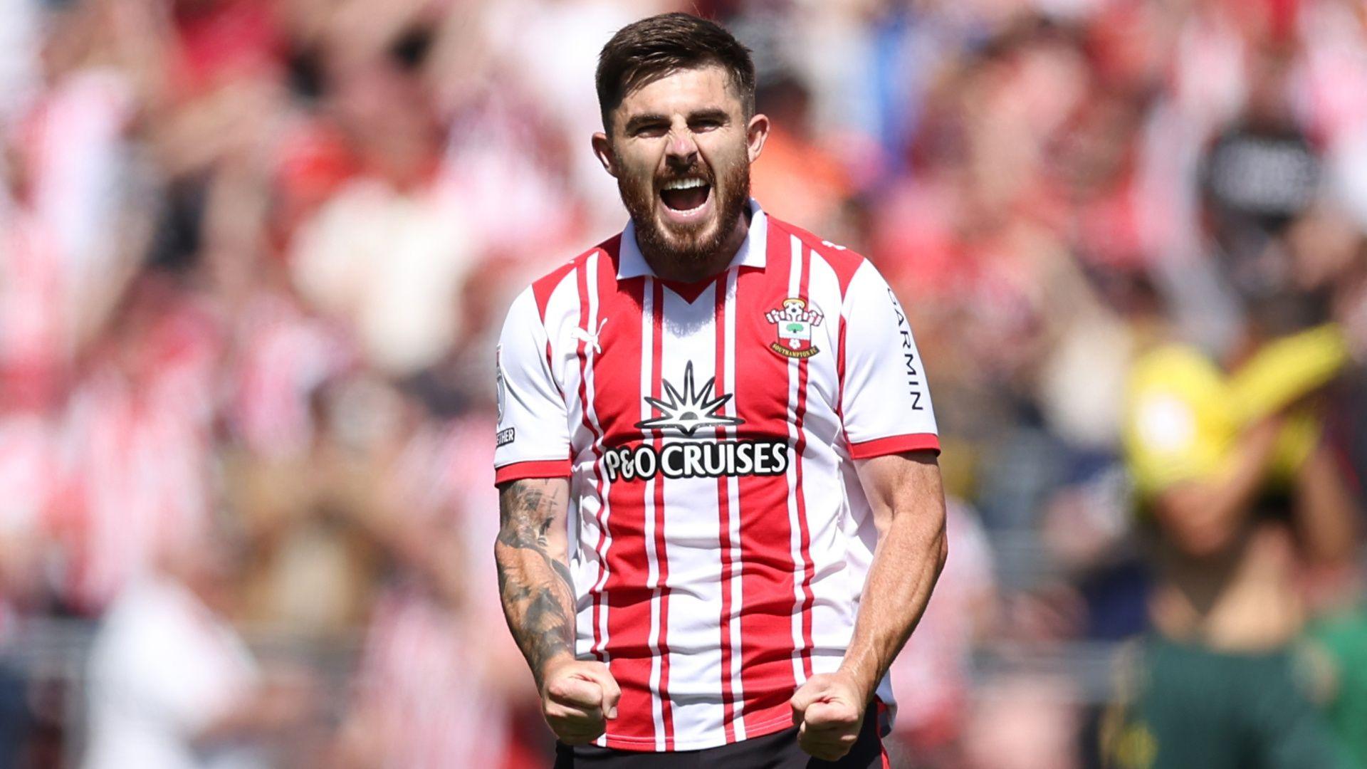 Ryan Manning opens his mouth wide shouting in celebration with a pleased look on his face and he holds his hands in fists in front of his waist. He wears a red and white vertically striped southampton shirt.
