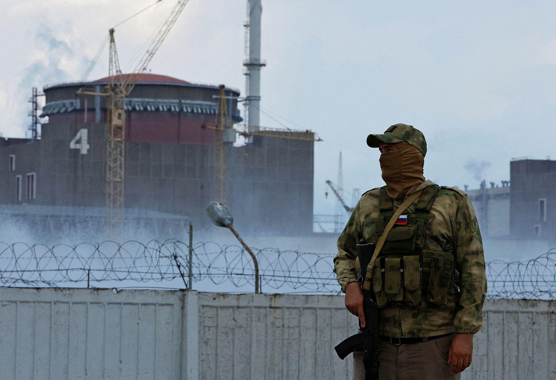 A serviceman with a Russian flag on his uniform, wearing military camouflage and a military baseball cap and balaclava stands guard with his hand on his weapon in front of a barbed wire fence with the Zaporizhzhia nuclear power plant in the background, in the Russian-controlled city of Enerhodar in the southern Zaporizhzhia region on 4 August 2022.