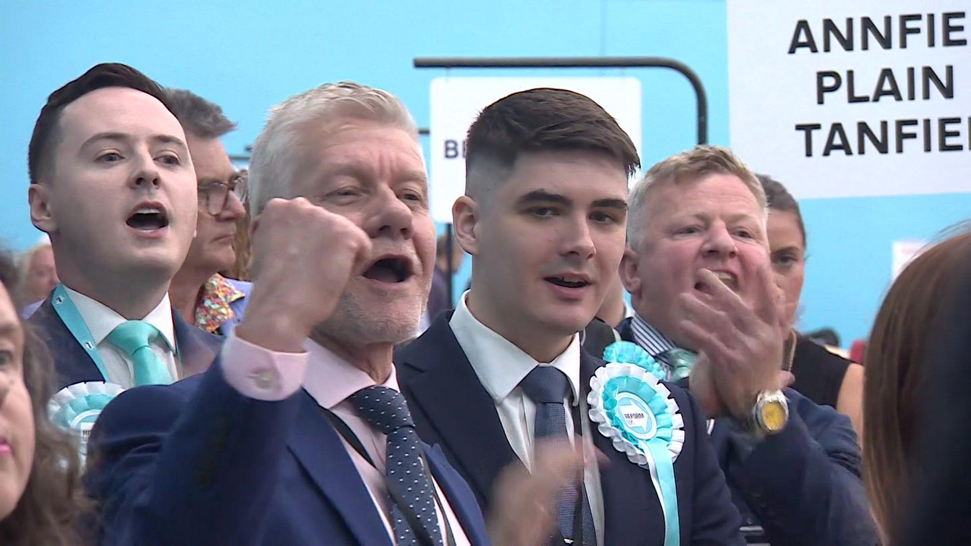 Reform UK celebrating. There are four men pictured, two are wearing rosettes.