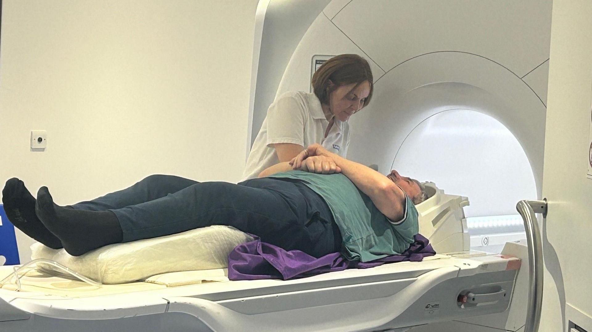 A man lying on an MRI bed scanner wearing a blue polo shirt and navy blue trousers