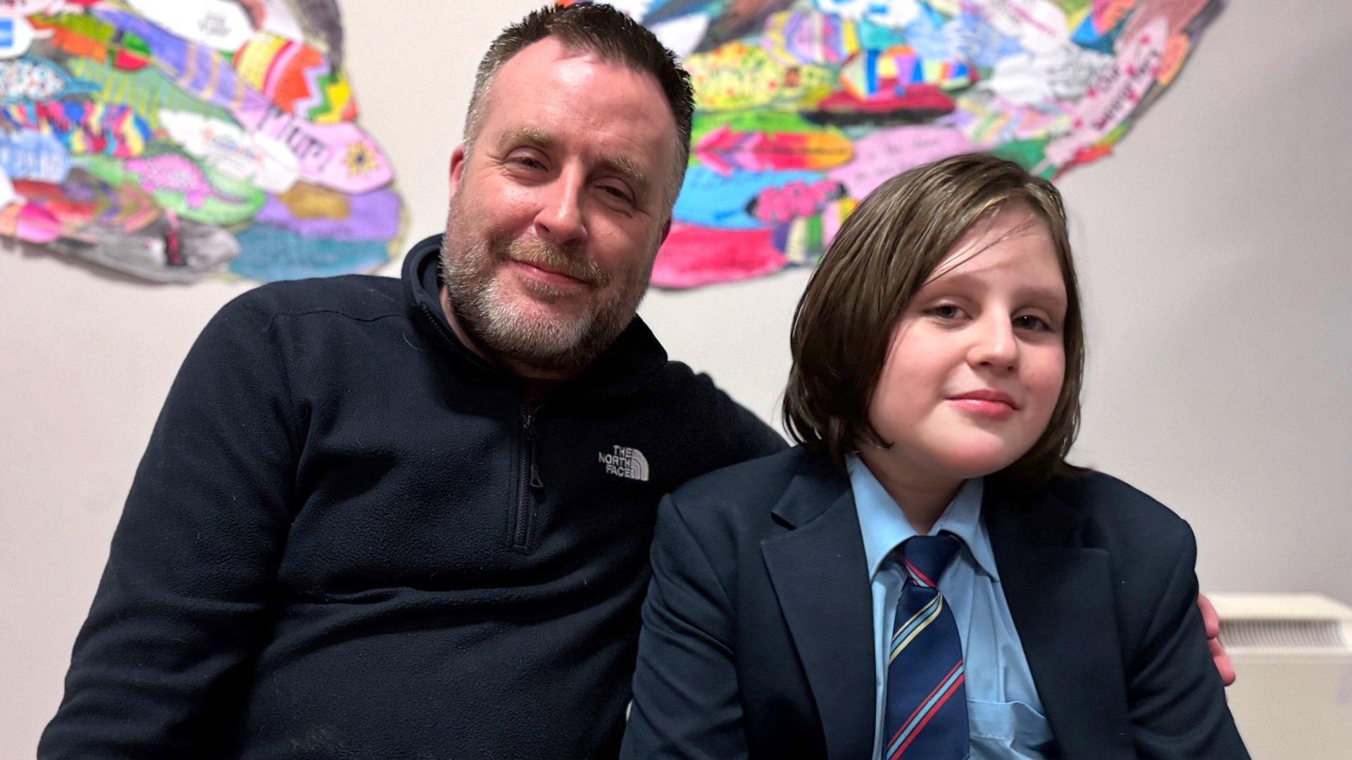 Cathal, in a dark North Face quarter-zip, has his arms around his son Ruairí who has long brown hair and is wearing a blue school blazer, light blue shirt and blue tie.