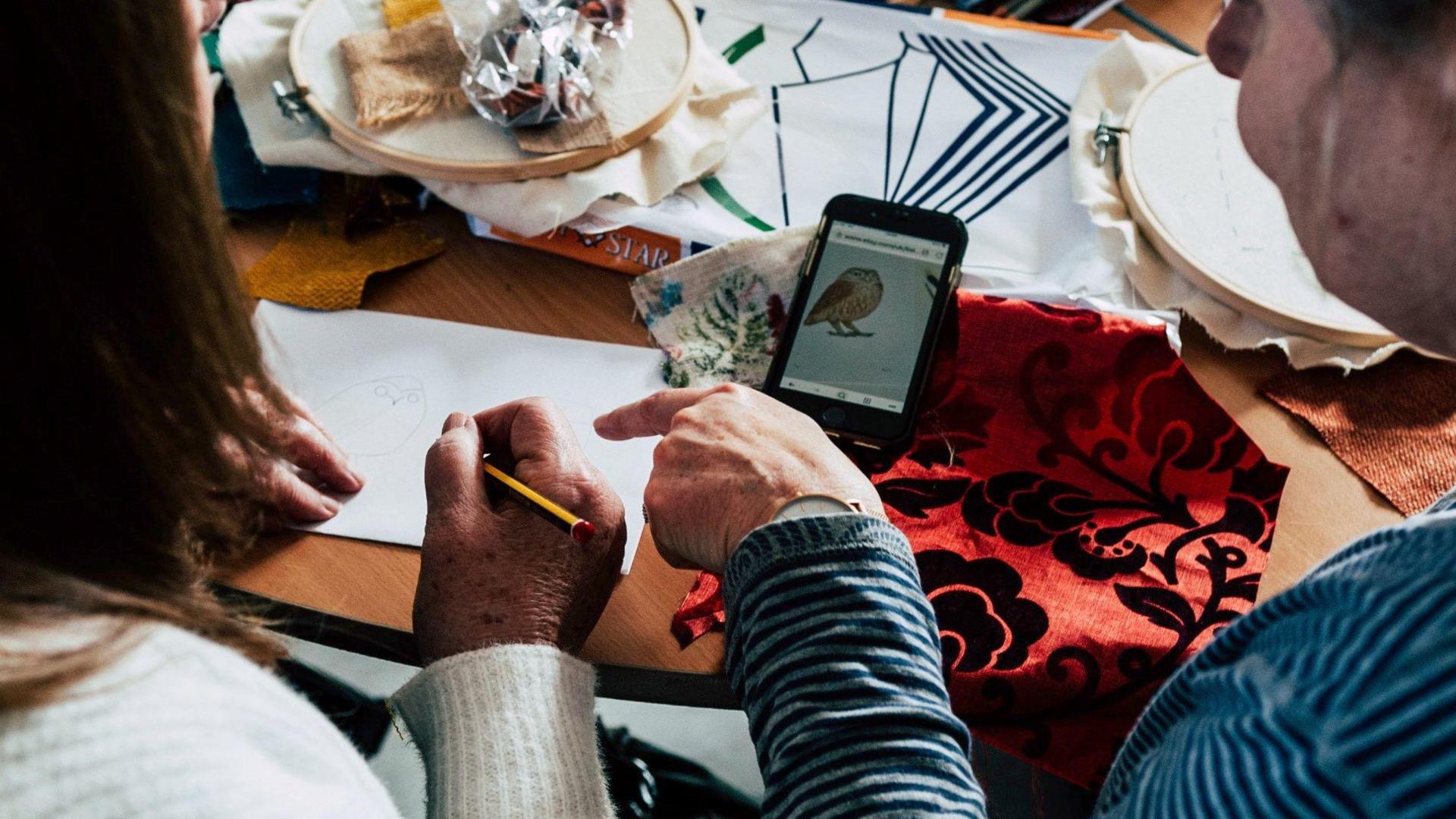A close up of two women sat next to each other pointing at a piece of paper. The woman on the left holds a pencil, there is a phone nearby with an illustration of an owl they are drawing. Pieces of fabric are scattered on the table. 