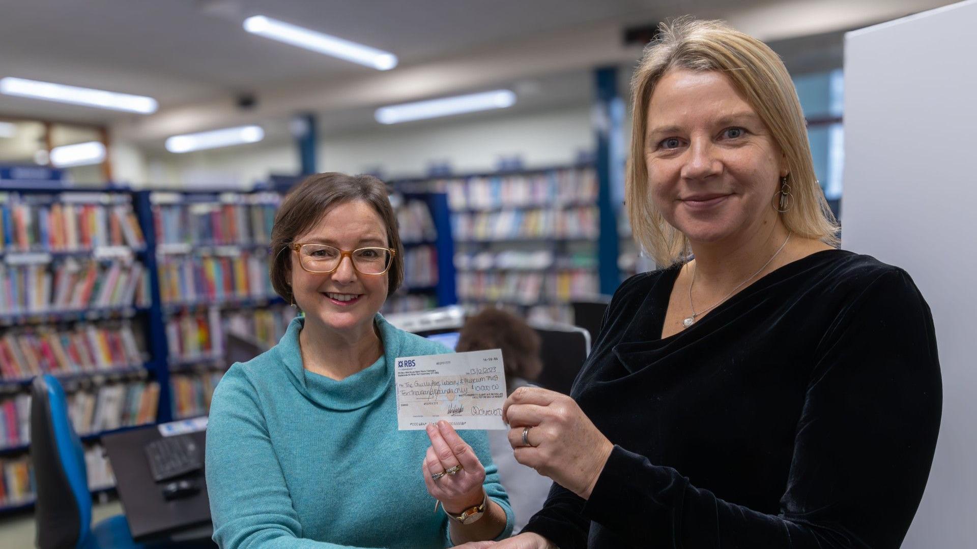 Becky Eker and Cornelia James pictured holding the cheque together. Ms Eker is wearing a light blue jumper and has short brown hair and brown glasses. Ms James, who is slightly taller, is seen wearing a black dress with blonde shoulder-length hair. They are both in the library with shelves of books behind them.