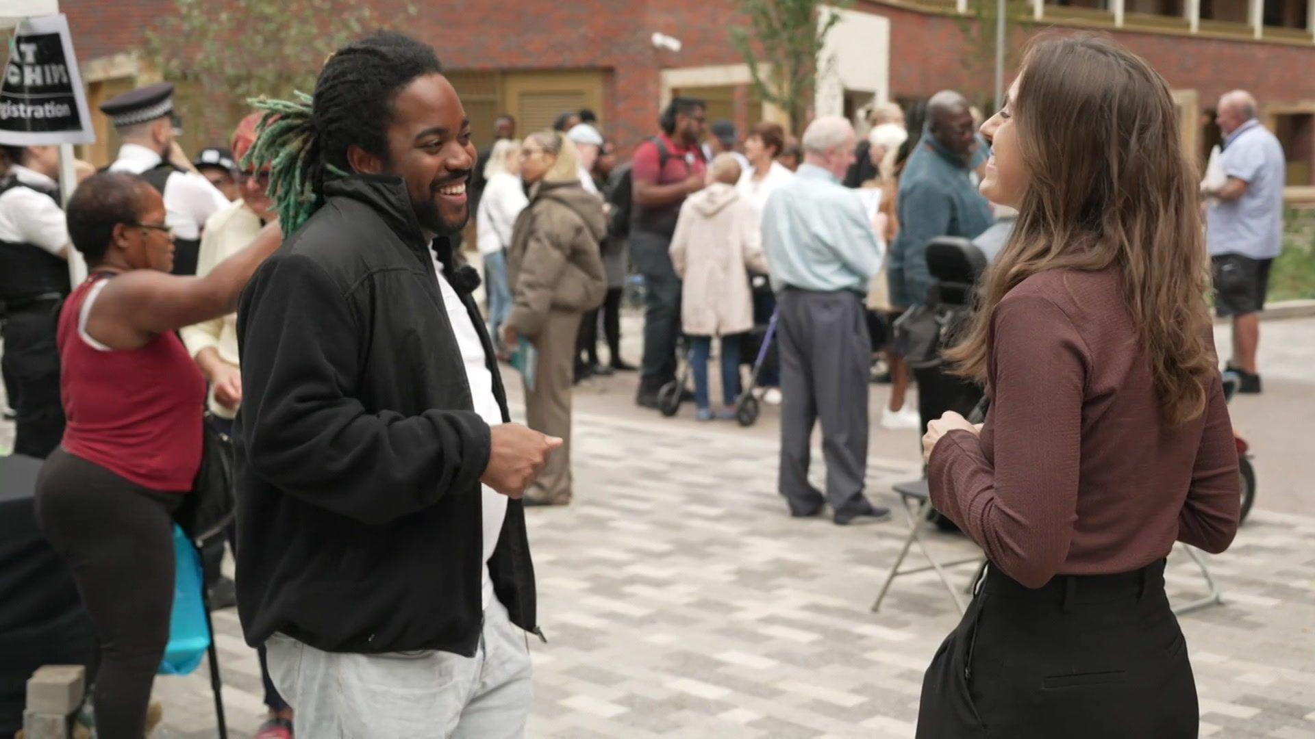 JJ standing among a group of people. He is wearing a black jacket over a white top and grey trousers