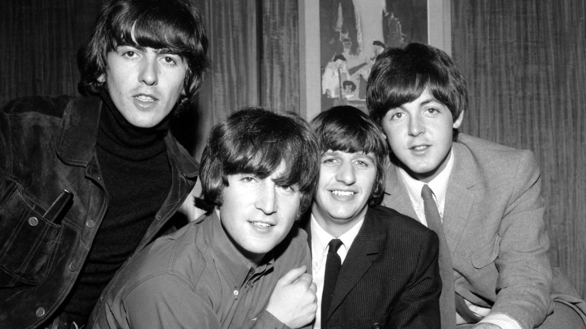 George Harrison, John Lennon, Ringo Starr and Paul McCartney, with mop-top haircuts and wearing smart casual clothes, pose for a black and white photo in an office