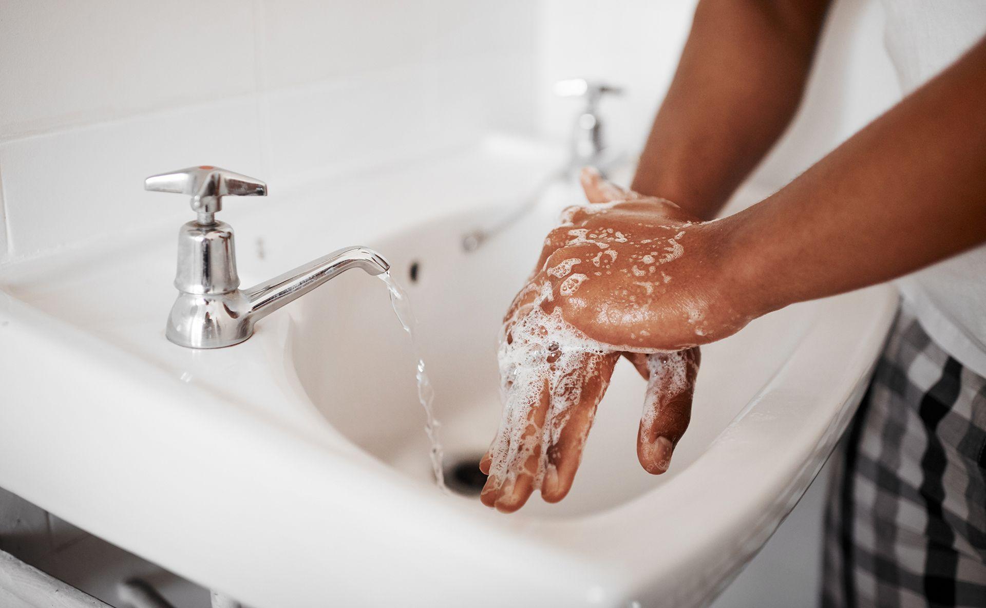 Close up on a woman's hands and lower arms as she washes her hands with soap under a running bathroom tap. The sink is ceramic white, and the woman is wearing a white shirt and striped trousers.