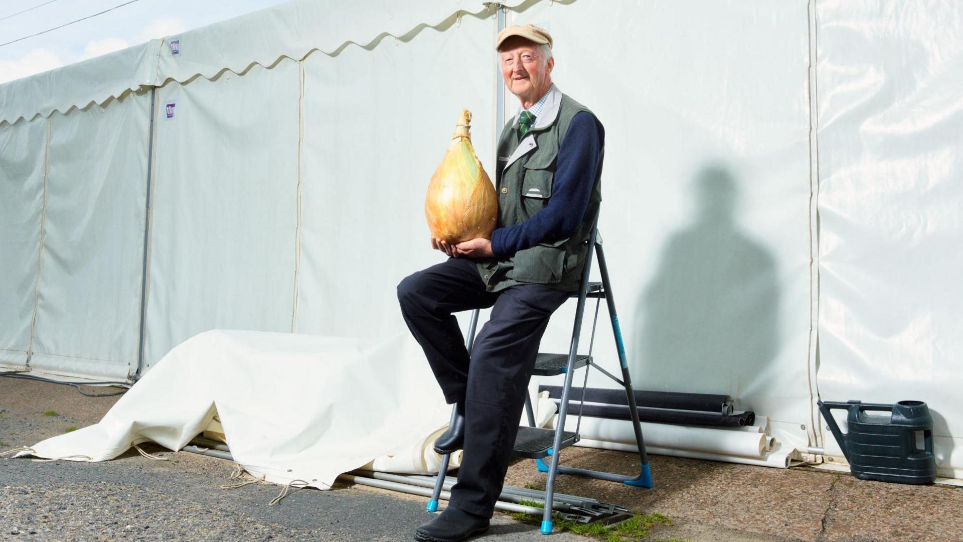 A man sitting on a step ladder outside a white tent. He is wearing a cap and a green sleevless jacket, dark trousers and jumper and a shirt and tie. in both hands he holds a heavy very large onion. 