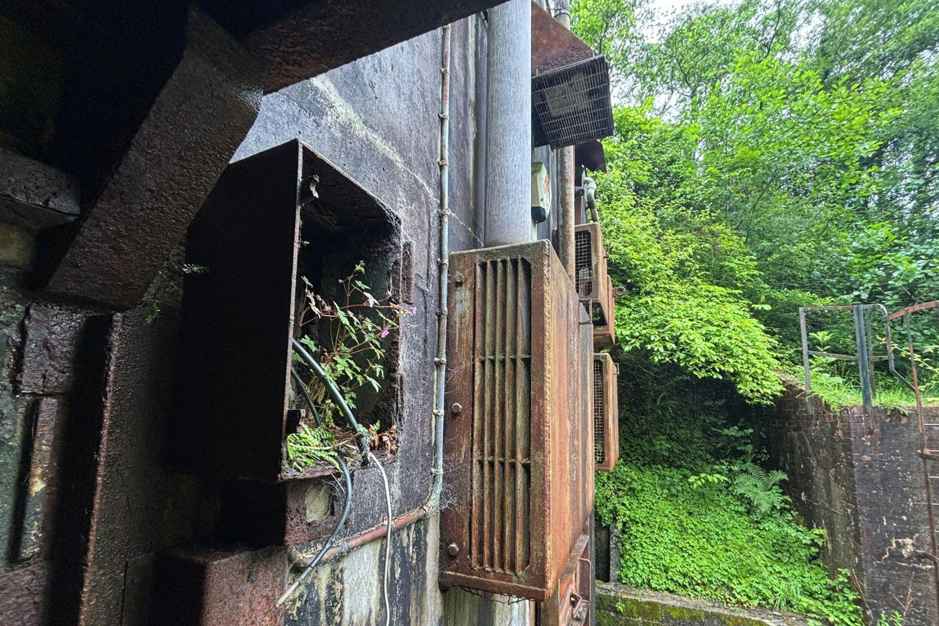 A rusty exterior of the bunker with wires protruding. The bunker looks overgrown.