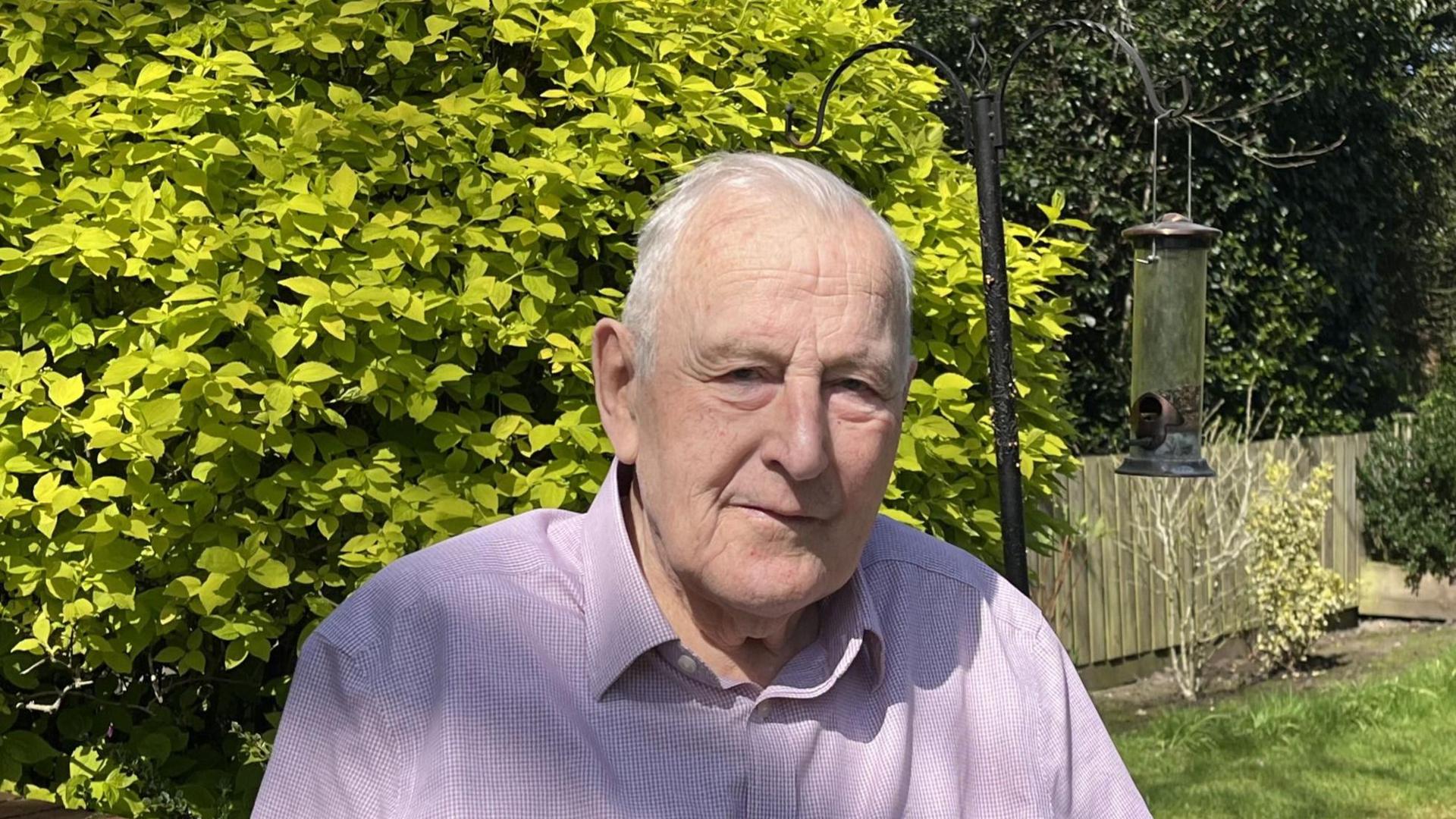 Peter Walker from Pewsey looks at the camera from his garden, a bird feeder and green hedge in the background. He wears a shirt.