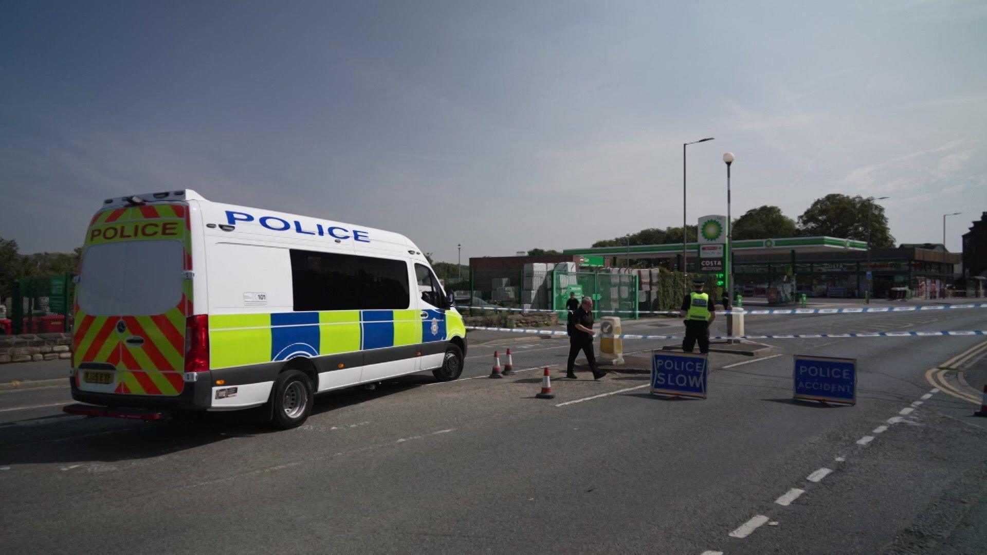 Police officers standing guard next to police tape, with a petrol garage in the background. A police van is also parked on the road.