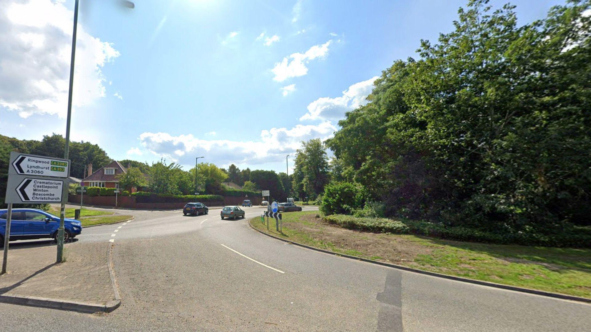 A view of Redhill roundabout. The centre of the roundabout can be seen to the right of the image, it is a round island covered in grass and decorated with some short hedging and a large tree. To the left, cars can be seen waiting to get onto the roundabout and others are seen in the distance driving around. There is a housing estate in the distance.