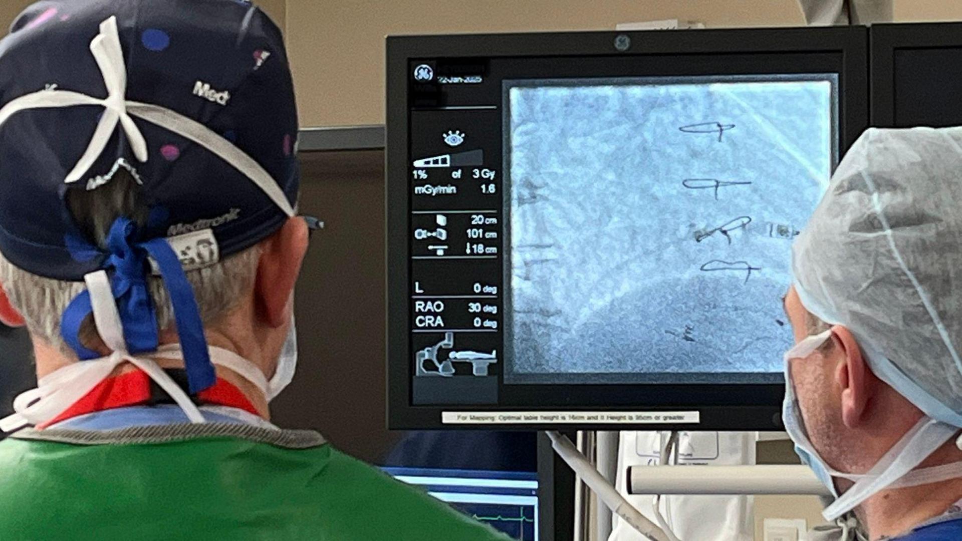 Two surgeons looking at an X-ray machine. They are both wearing surgical clothing and masks.