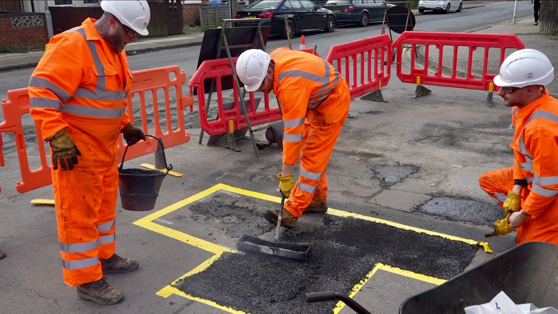 Highways crews are seen filling in a pothole on a road in Ipswich. The road can be seen cordoned off with red barriers while the crews are seen placing a material on the road surface. The area of the road they are working on has been marked with a yellow boundary. The crews are wearing orange high vis jackets and white hard hats. 