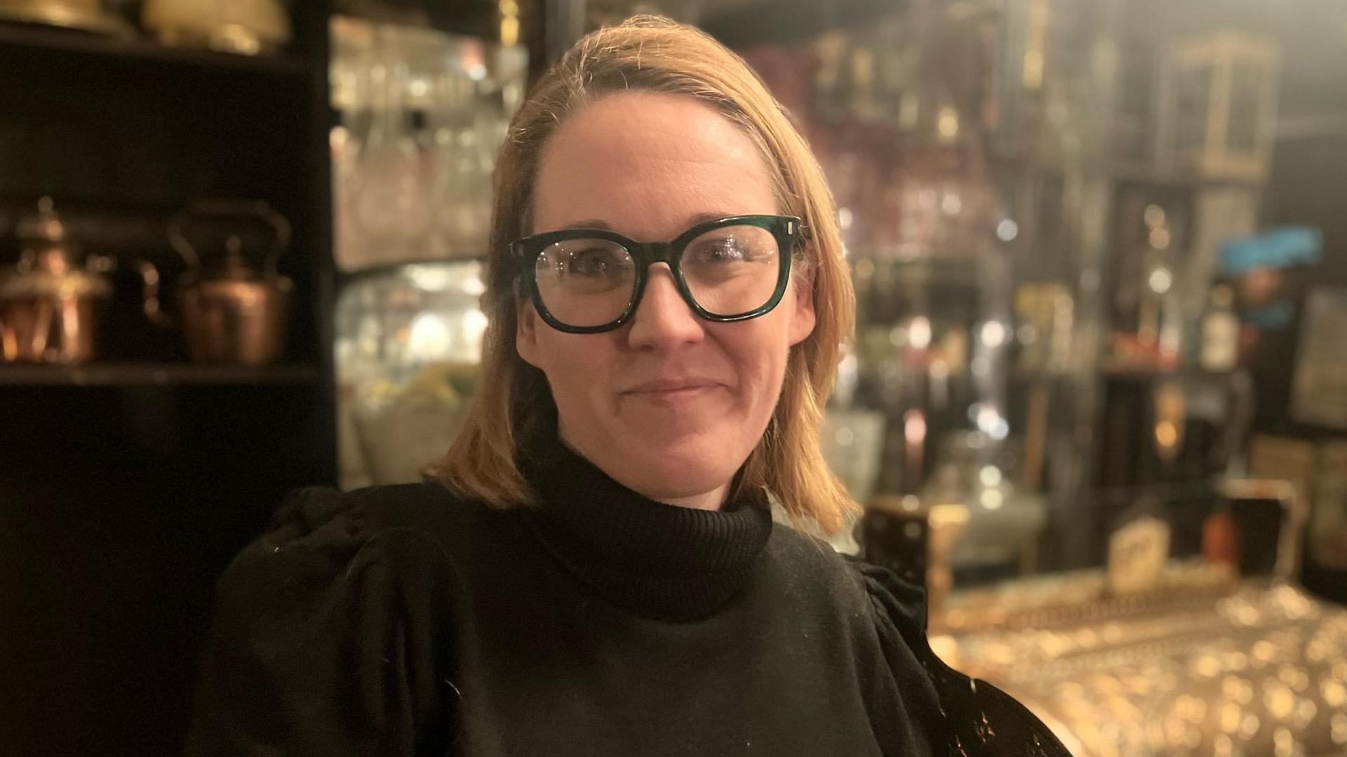 A blonde woman, wearing glasses, stands in a shopfront full of copper kitchenware.