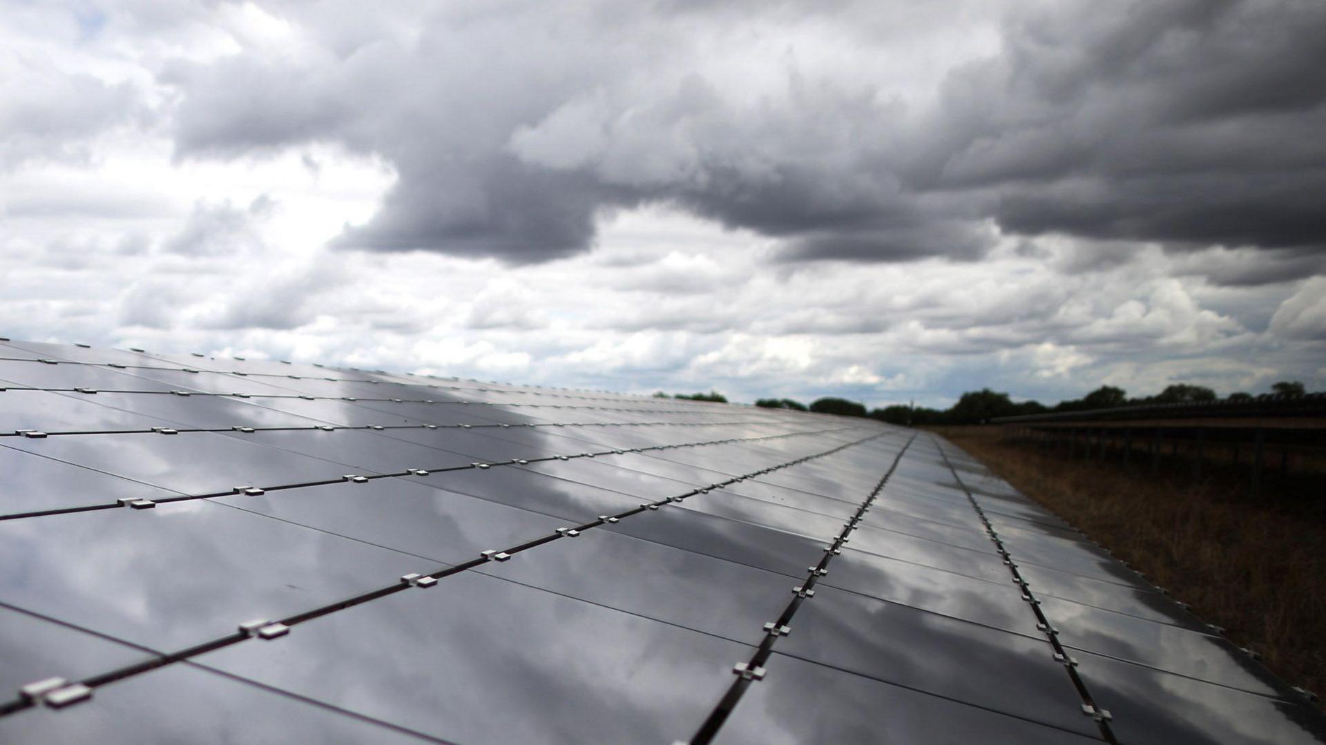 View of solar panels stretching into the distance in a field. The sky is cloudy and the clouds are reflected in the panels.