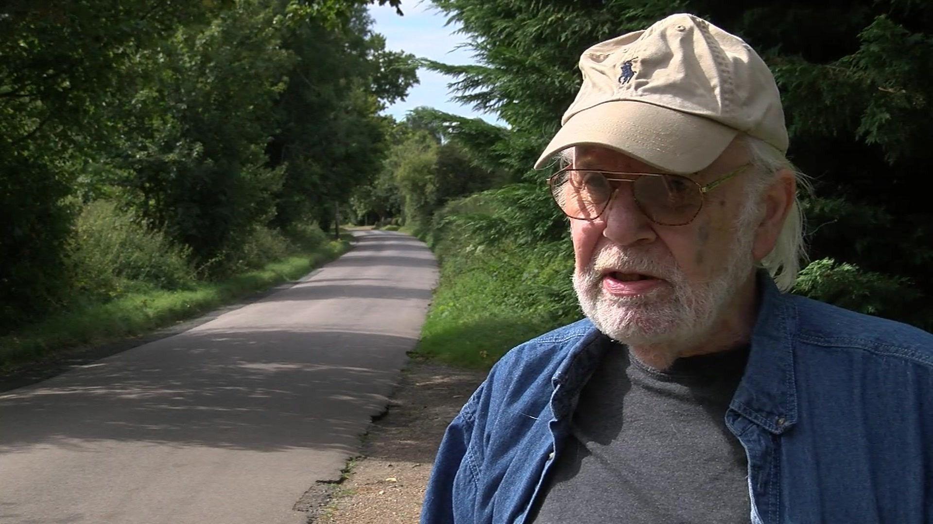 A man wearing glasses, a beige baseball cap, a dark grey t-shirt and a blue denim shirt over the top, standing in front of a narrow, single-carriageway road with shrubbery on both sides and no pavement.