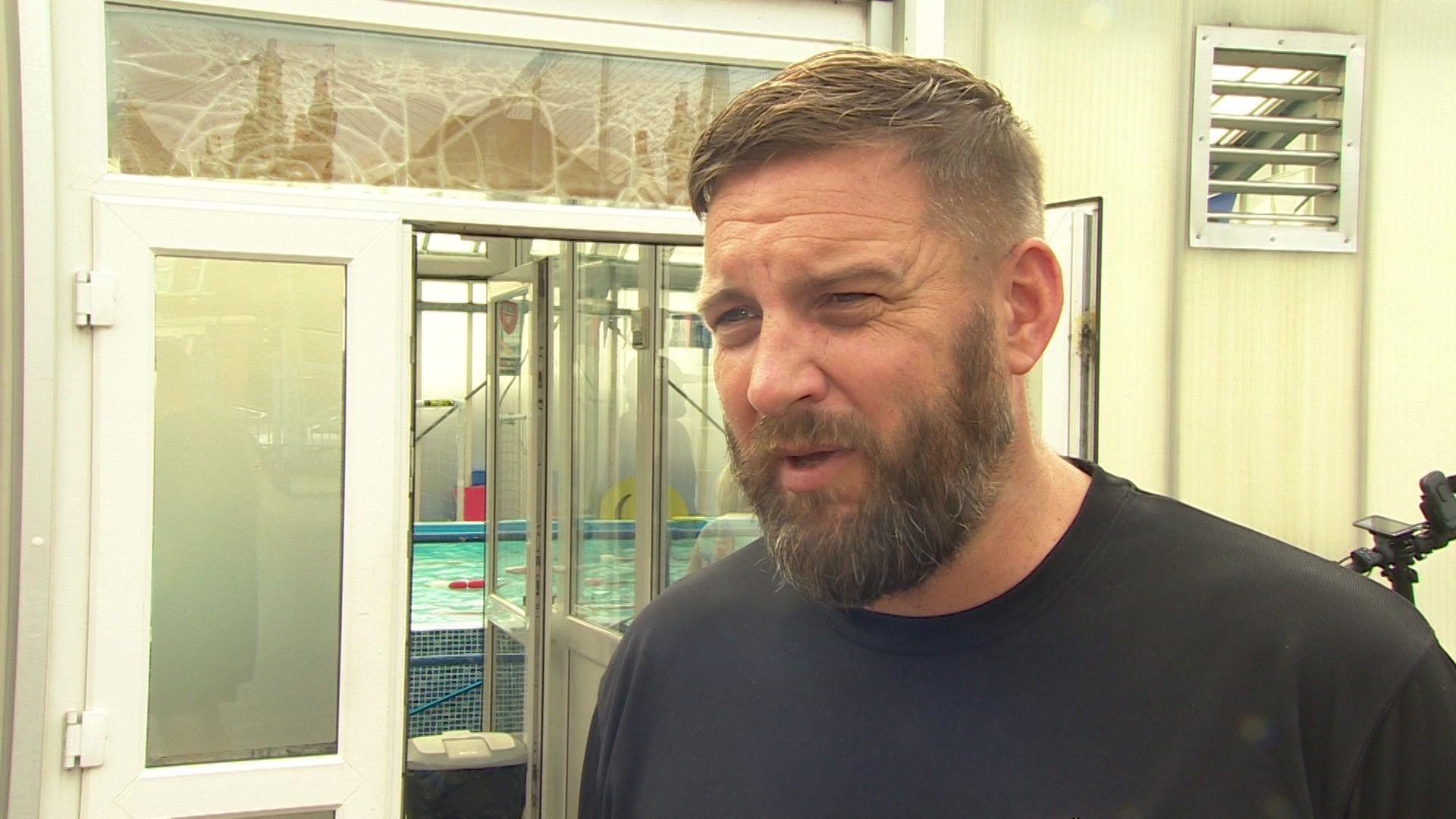 A white man stands outside a swimming pool looking off to the side of the camera talking.