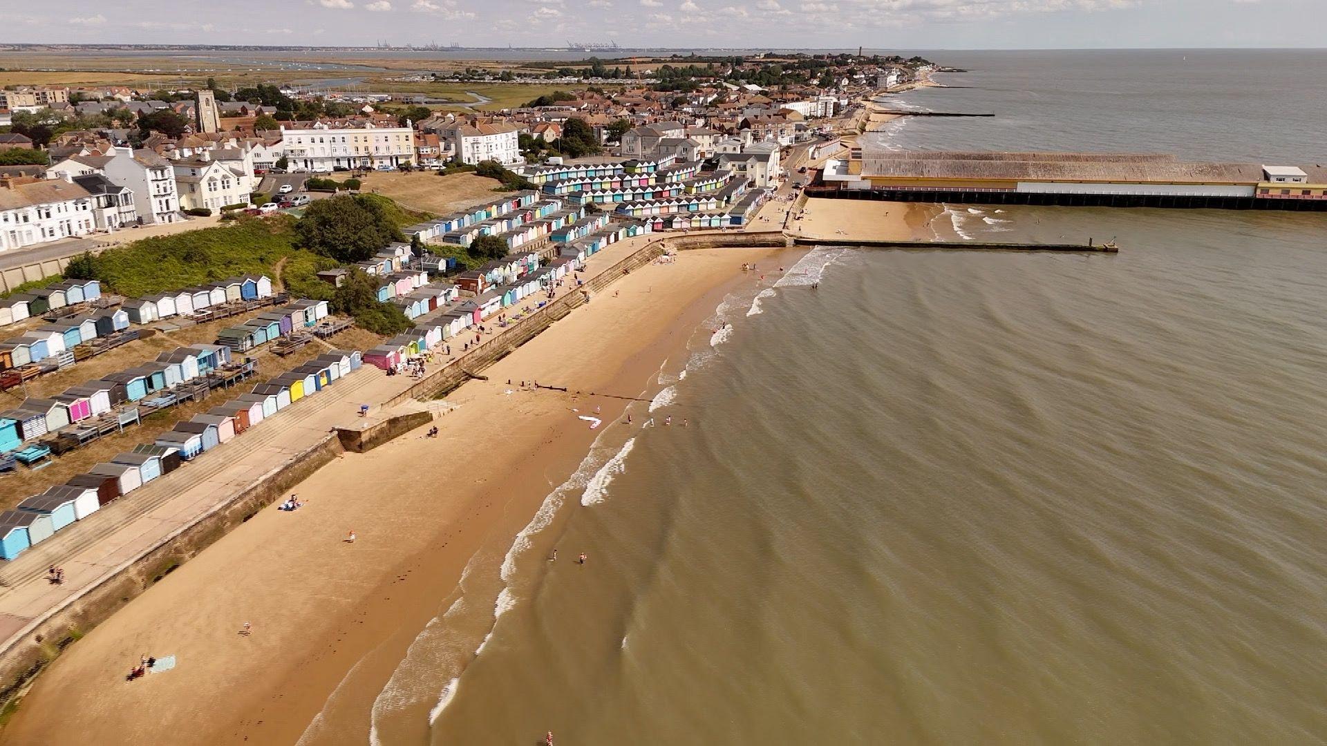 An aerial shot of the sea with a beach and pier. There is a small town on a hill and multi-coloured beach huts.