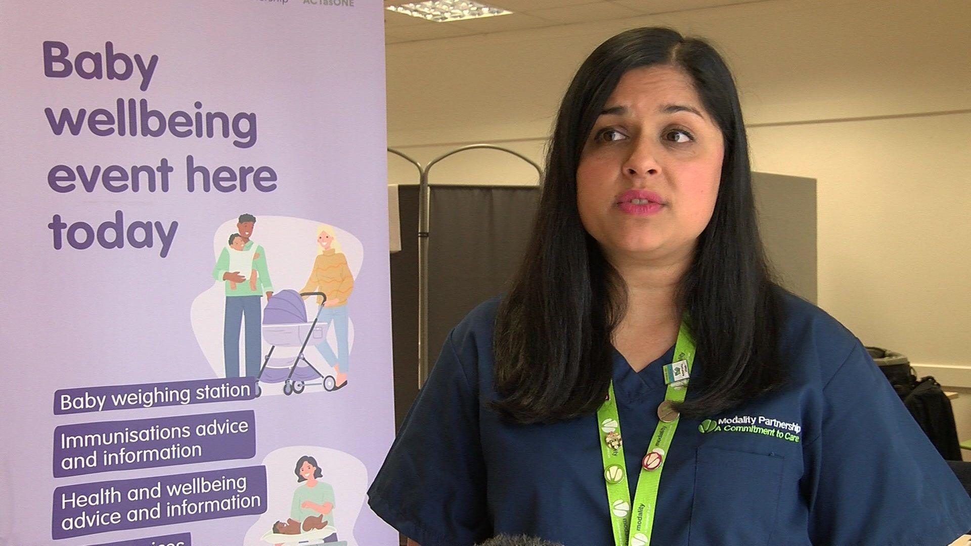 A woman in dark blue medical uniform stands next to a banner promoting a baby wellbeing event. 