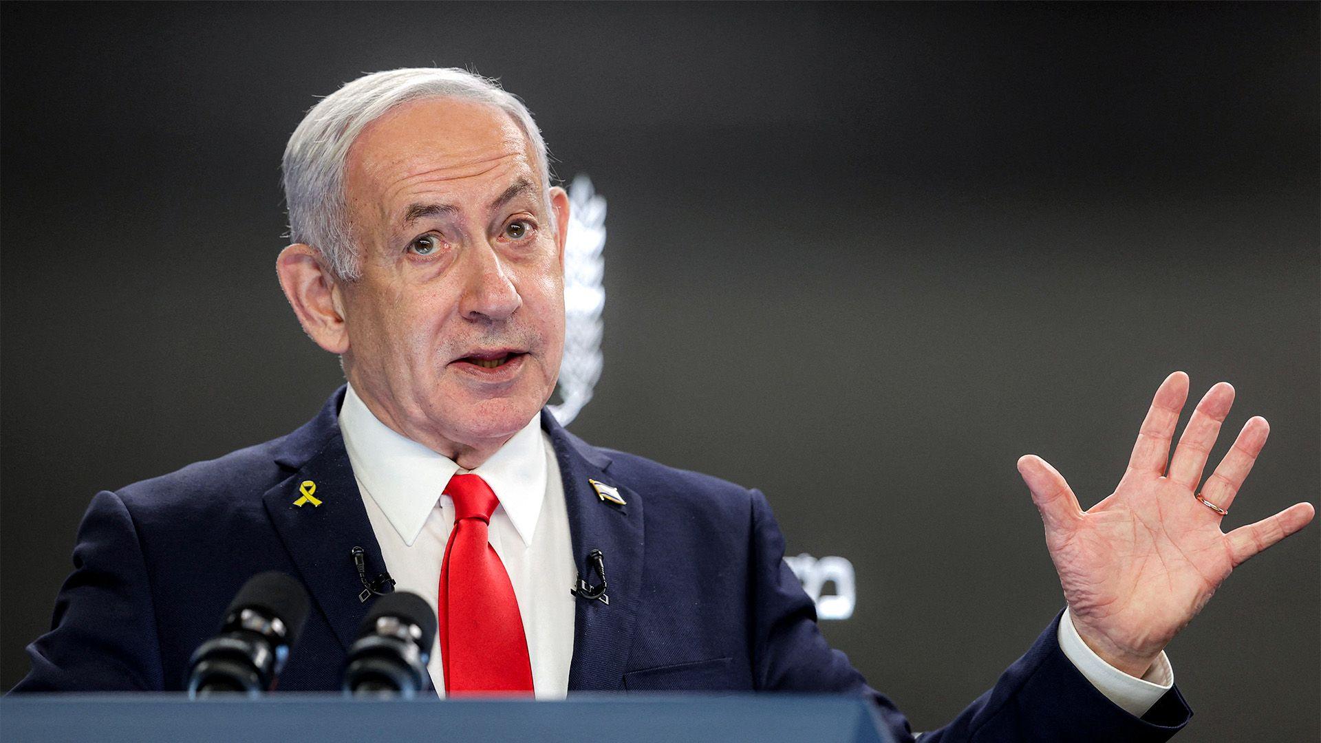 Israeli Prime Minister Benjamin Netanyahu speaks at a podium with microphones in front of him. He wears a dark suit, white shirt, and red tie, with a yellow ribbon pin on his left lapel. His right hand is raised with fingers spread apart.
