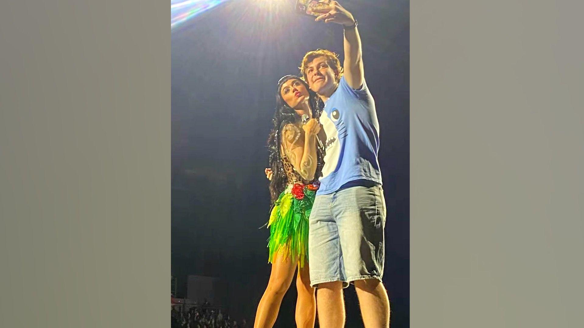 Ben, who is wearing a blue t-shirt with blue denim shorts, holds his phone up and takes a selfie with Katy Perry. She is holding a microphone and is wearing an animal print top and bright green skirt.