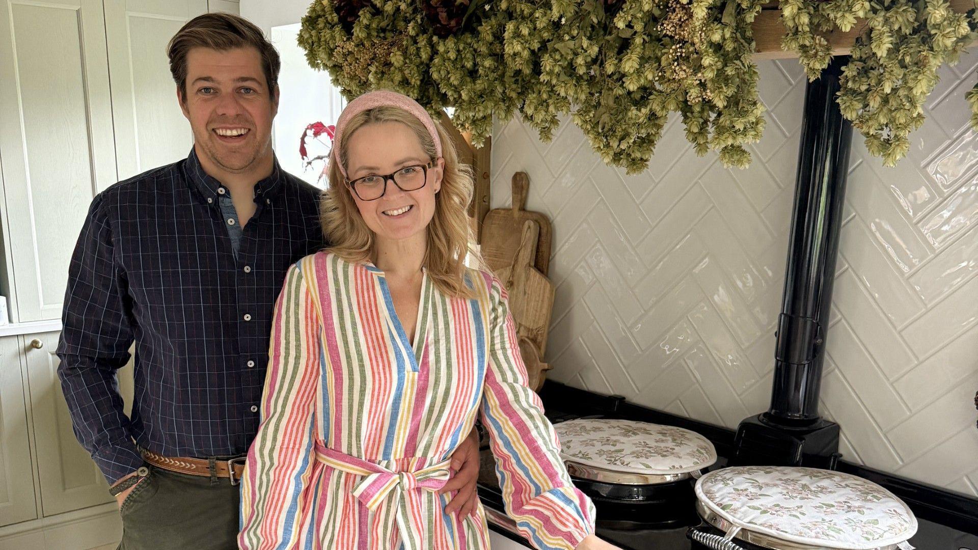 Aimee and Sean, a married couple, stand in front of an Aga unit. Aimee is wearing a striped dress in several colours and Sean is wearing a navy check shirt