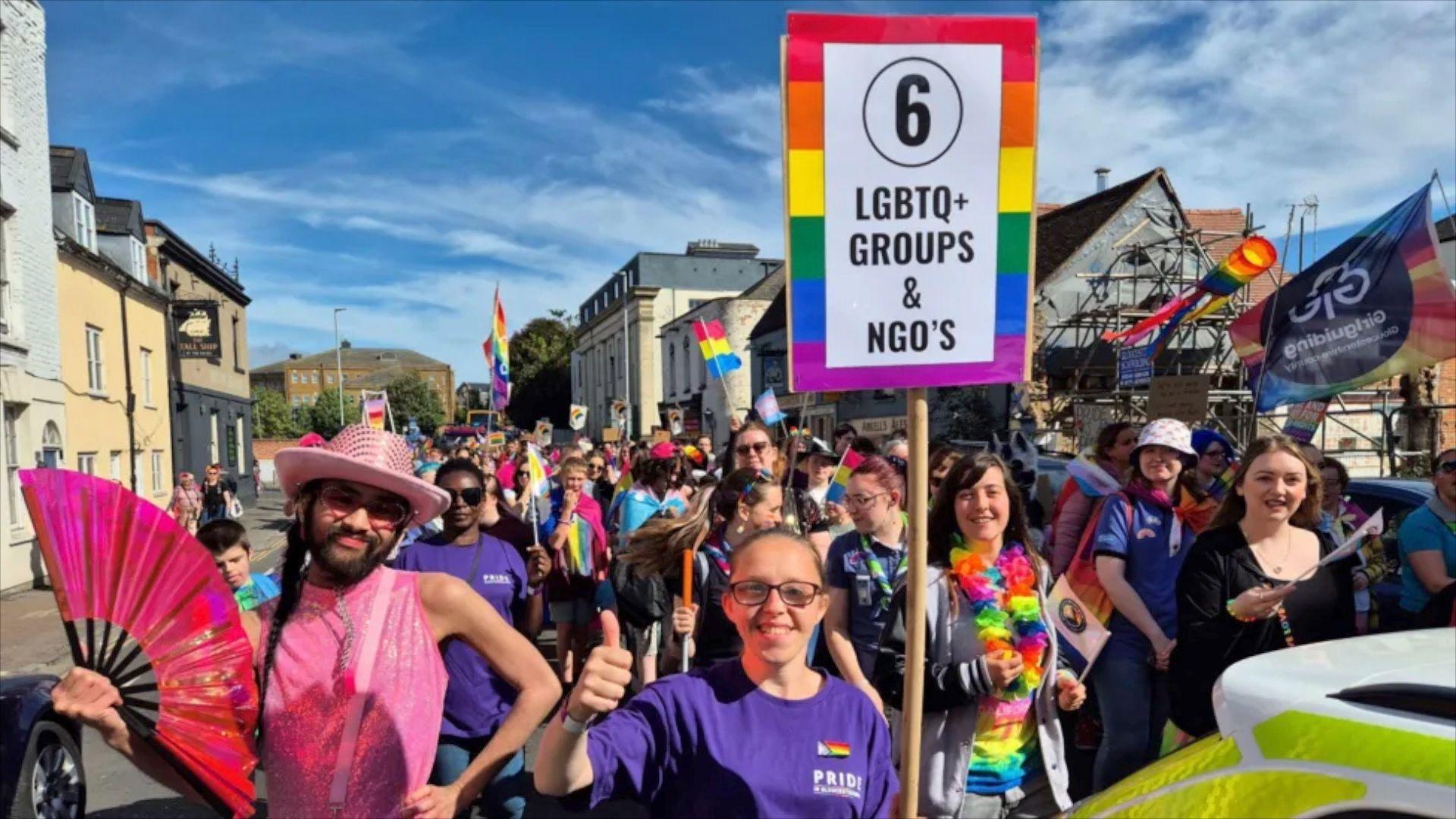 A group of people at a pride event wearing brightly coloured clothes with placards and flags. 