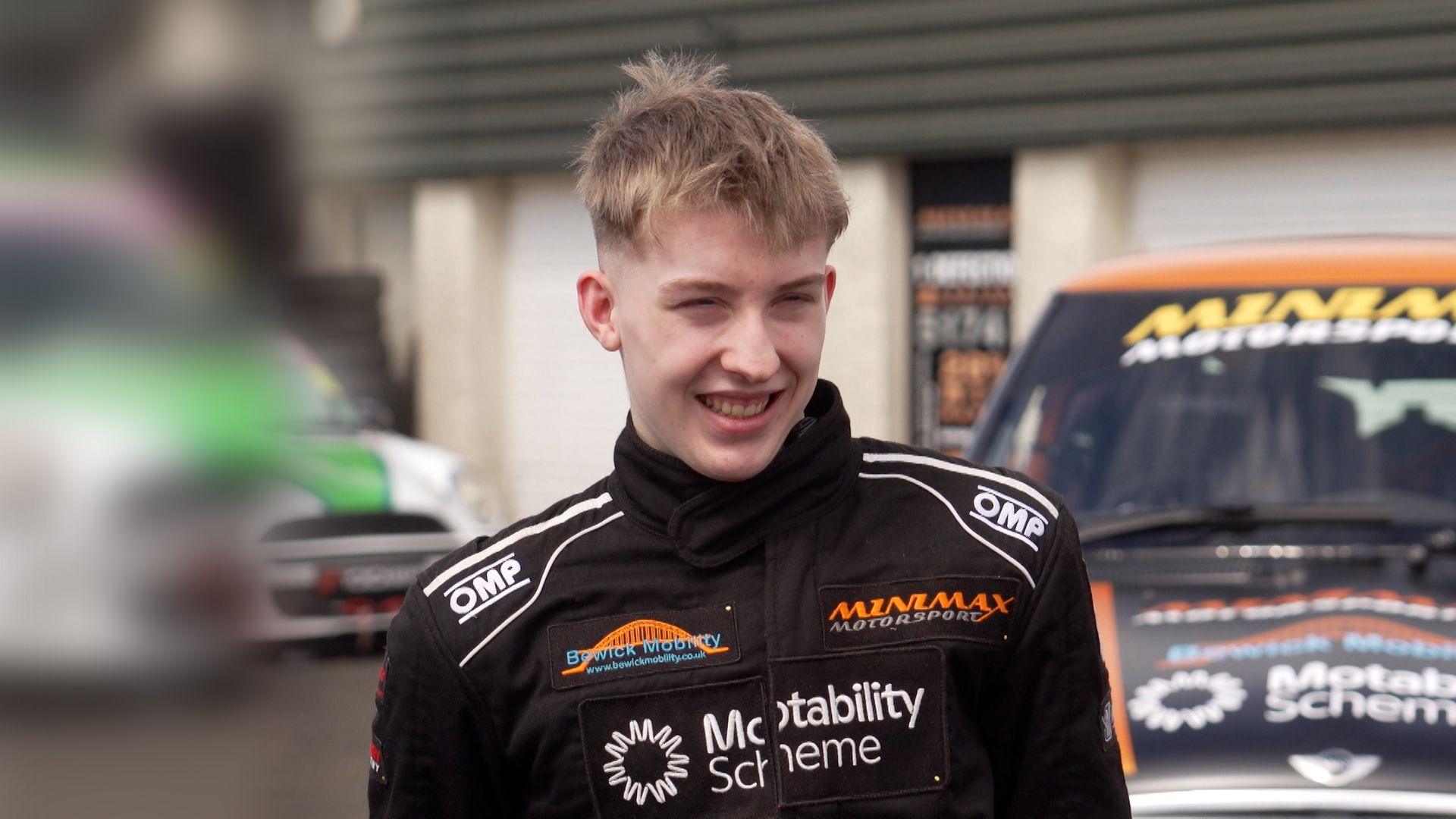Ally Chalmers, a 17-year-old boy with short, blond hair. He is looking off to the side and is smiling. He is wearing a black race suit with the names of sponsors on it. There are racing cars behind him.