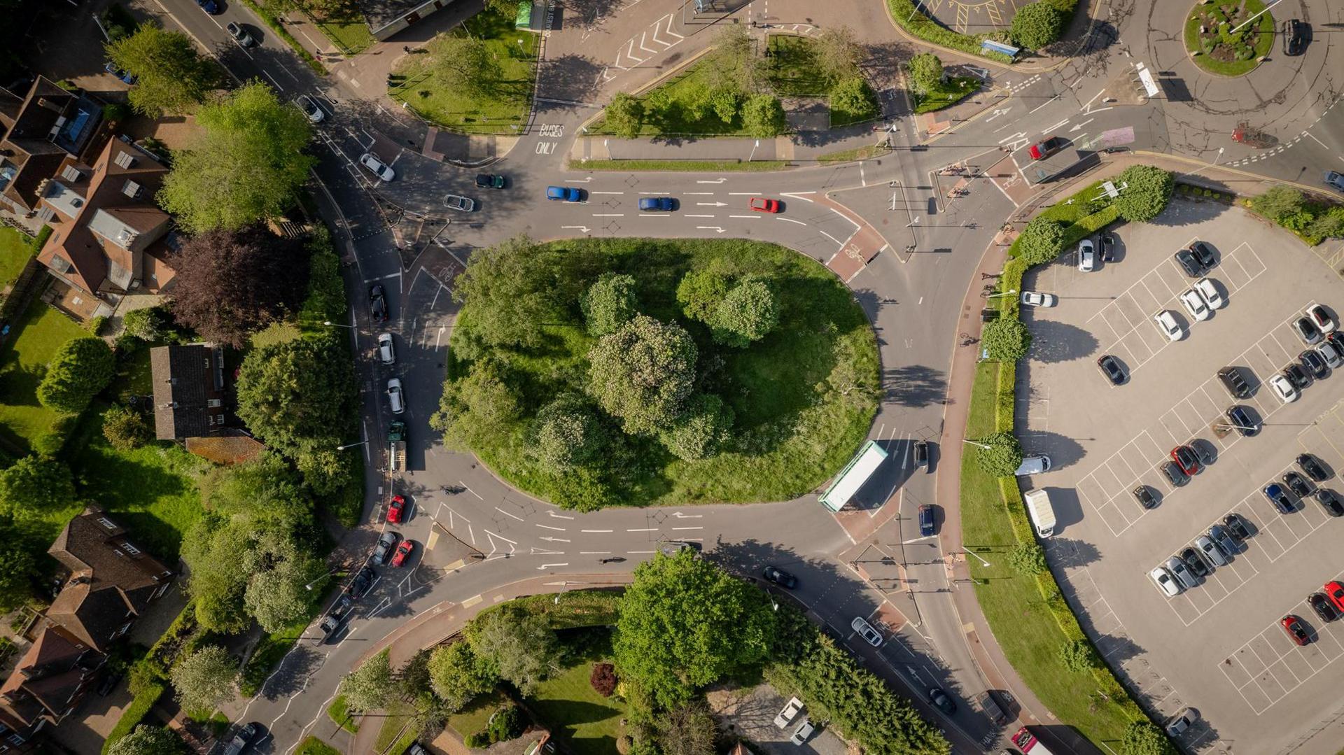 An aerial view of a roundabout with four exits and green bushes and trees in the centre.